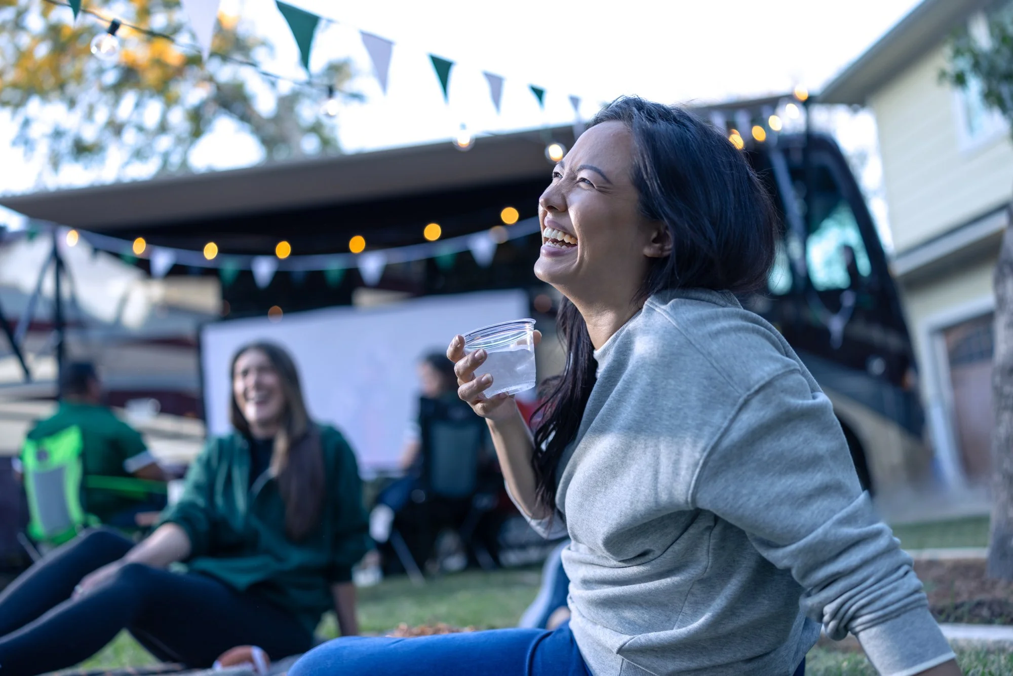 A woman laughing and smiling while sitting outdoors at a social gathering, holding a clear plastic cup with a drink. There are string lights and bunting decorations in the background, with other people seated nearby, suggesting a festive event.