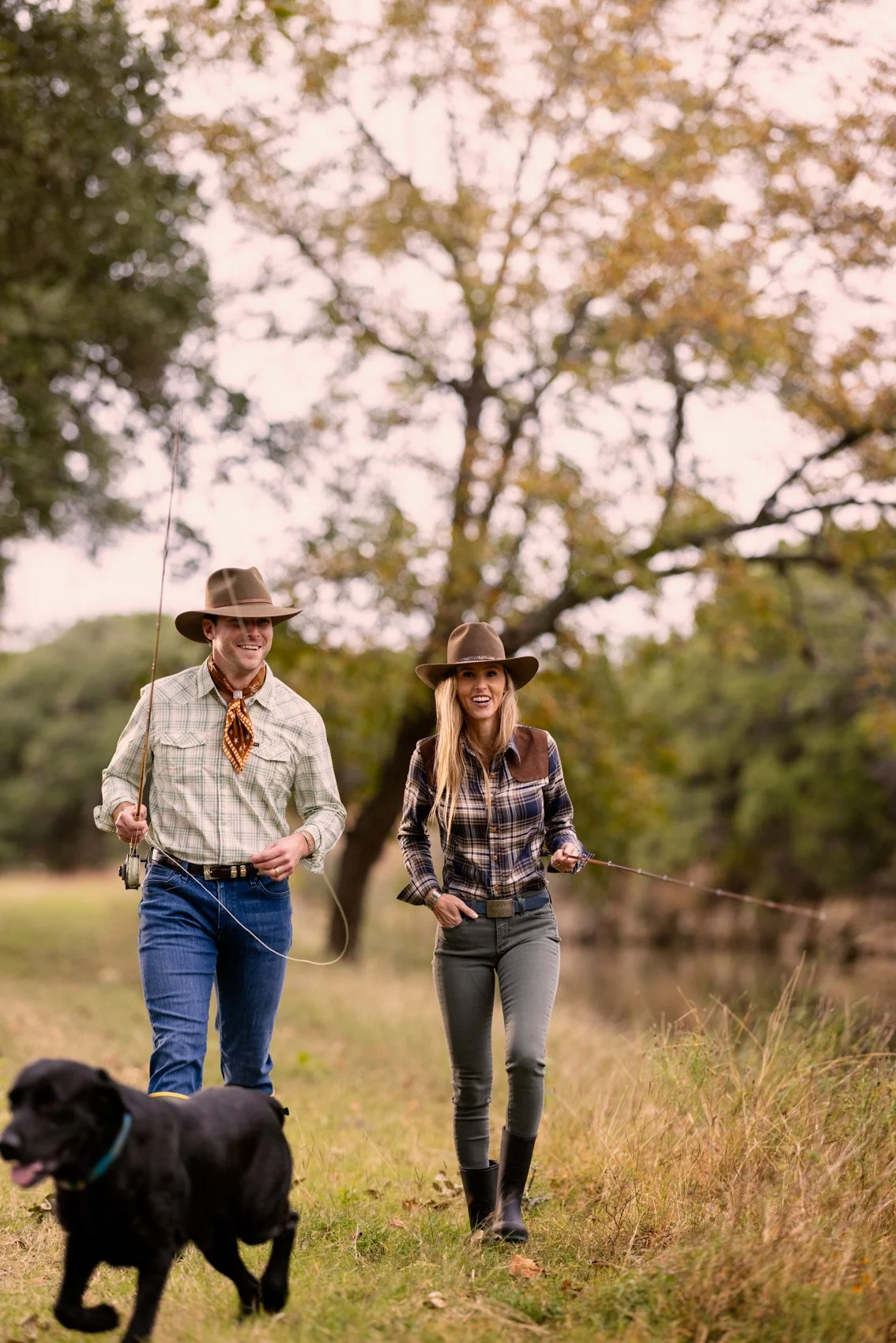 A man and woman dressed as cowboys walking outdoors on a grassy trail with a black dog. The man is holding a fishing rod, and they are both smiling. The trees surrounding them have autumn leaves, and the sky is cloudy.