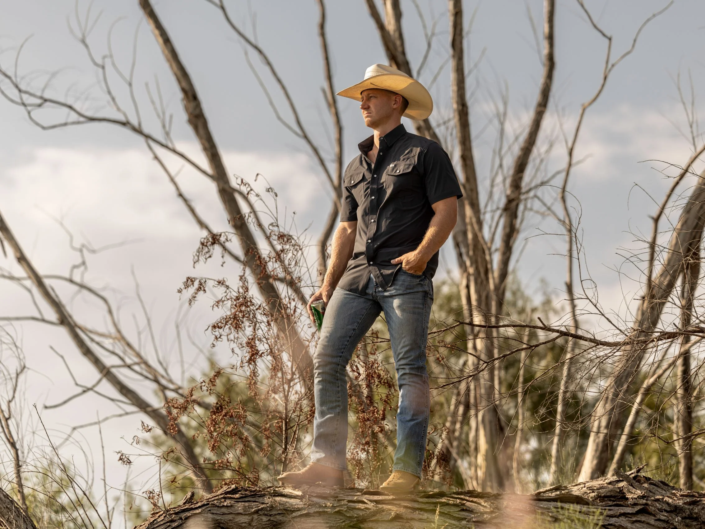 A man wearing a cowboy hat and black shirt stands on a fallen tree branch in a wooded area with leafless trees and cloudy sky.