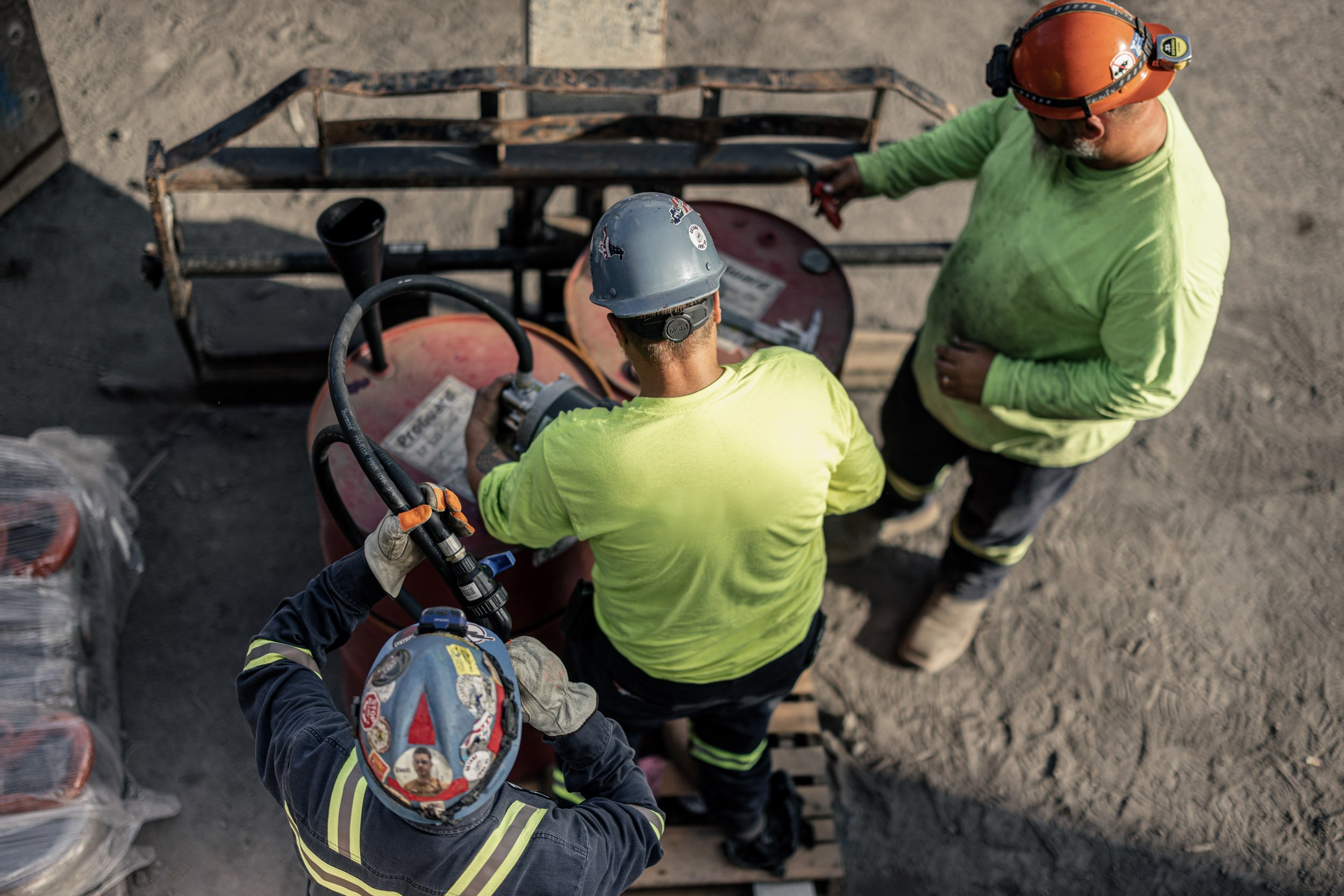 Two construction workers wearing safety helmets and bright yellow-green shirts working with a piece of equipment outdoors on a construction site.