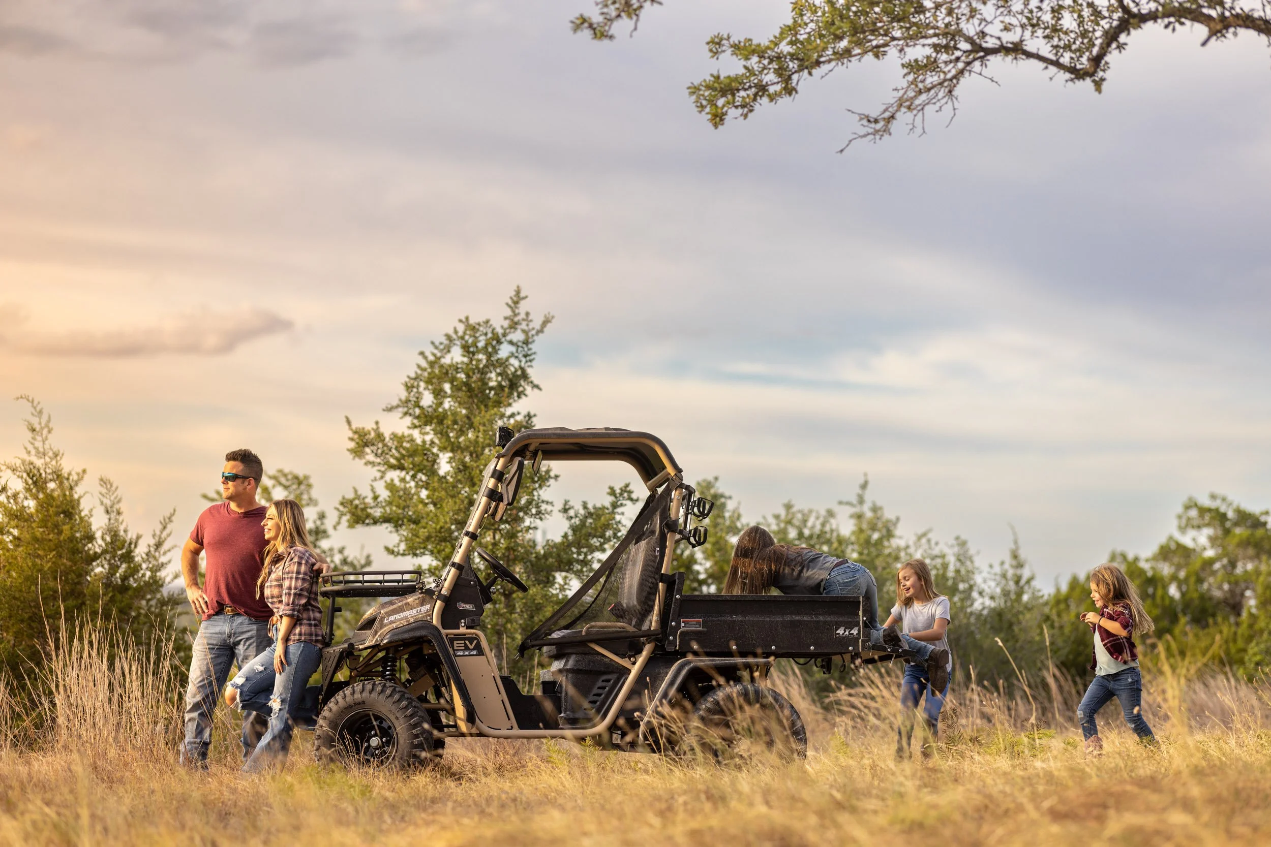 Groups of people, including adults and children, gather outdoors around a utility vehicle in a grassy field with trees and a cloudy sky in the background.