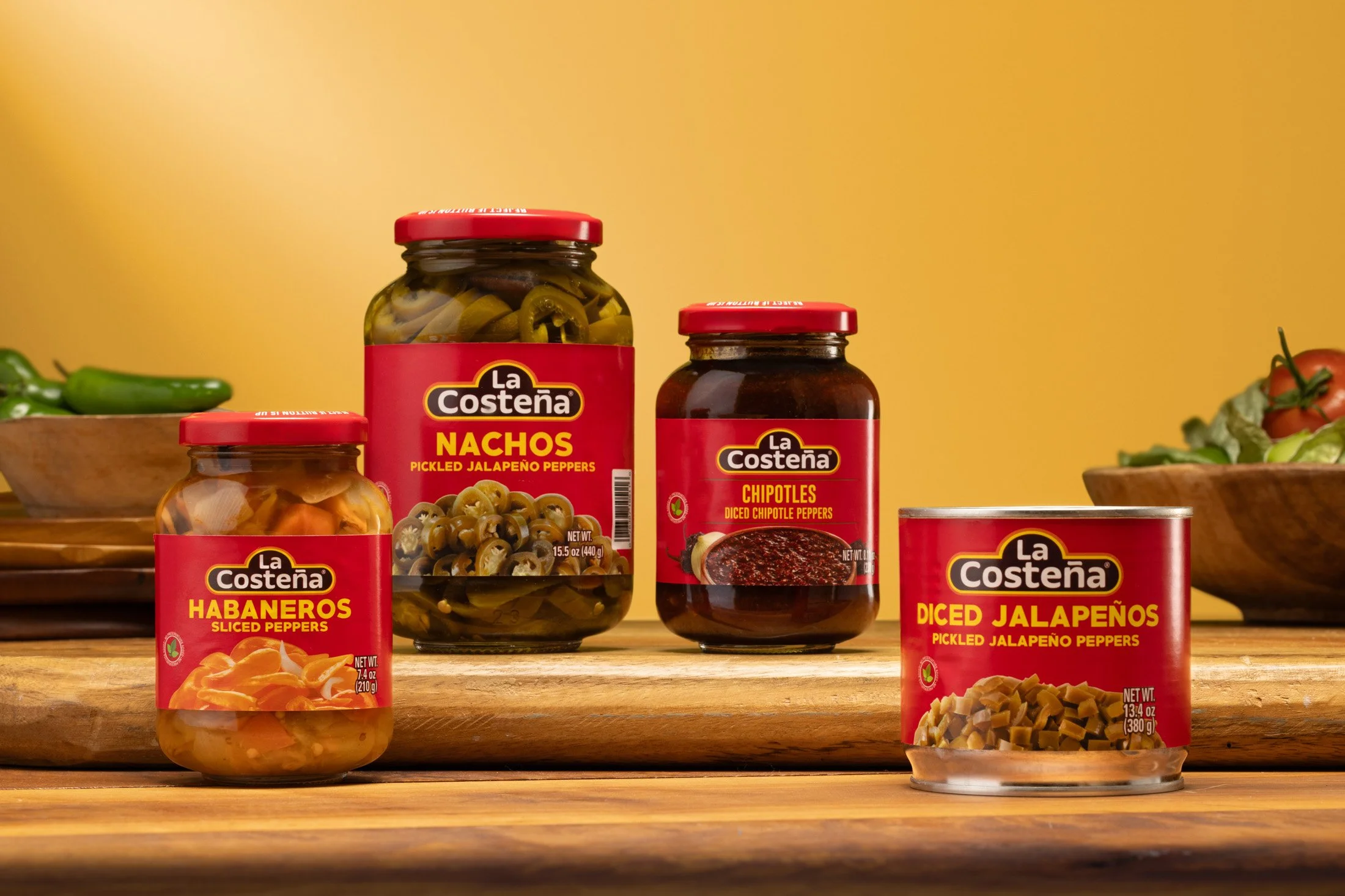 Various jars of La Costeña brand canned peppers, including sliced habaneros, pickled jalapeños, diced chipotle peppers, and pickled jalapeños, placed on a wooden surface with a yellow background.