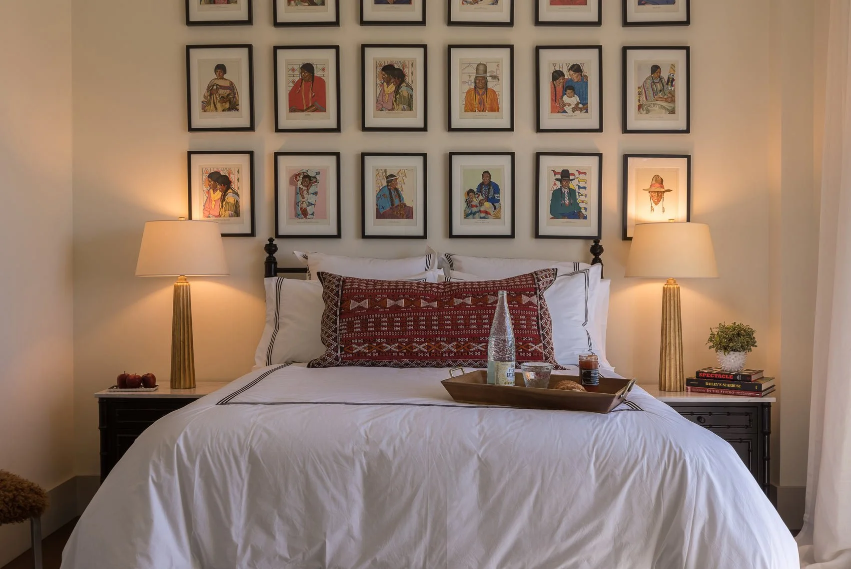 A neatly made bed with white linens and decorative pillows, flanked by two nightstands with lamps. Behind the bed, a wall decorated with framed artwork features portraits of Indigenous people. On the bed, a tray holds a bottle of water, a glass, a sm