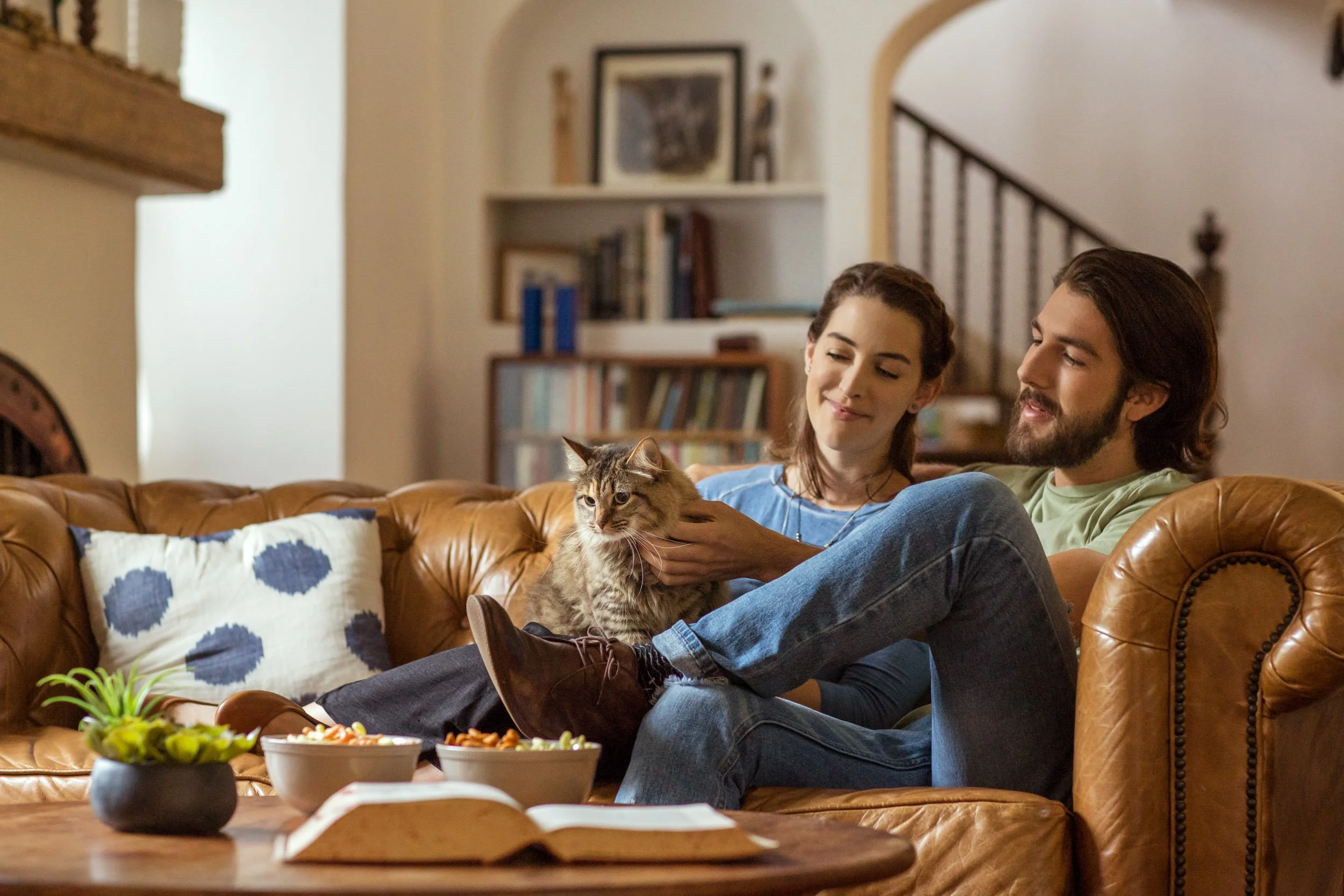A young woman and a young man sit together on a brown leather sofa in a living room, smiling and holding a tabby cat. The room has a bookshelf with framed art and a staircase in the background. A table with bowls of snacks is visible in the foregroun