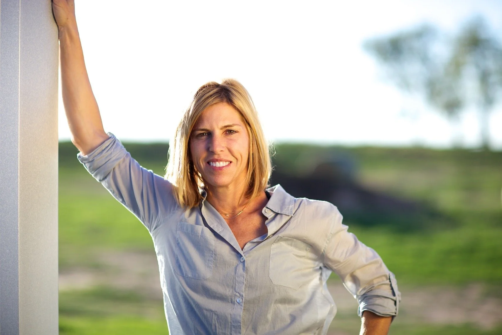Woman with blonde hair wearing a light-colored button-up shirt, standing outdoors with greenery in the background, smiling slightly.