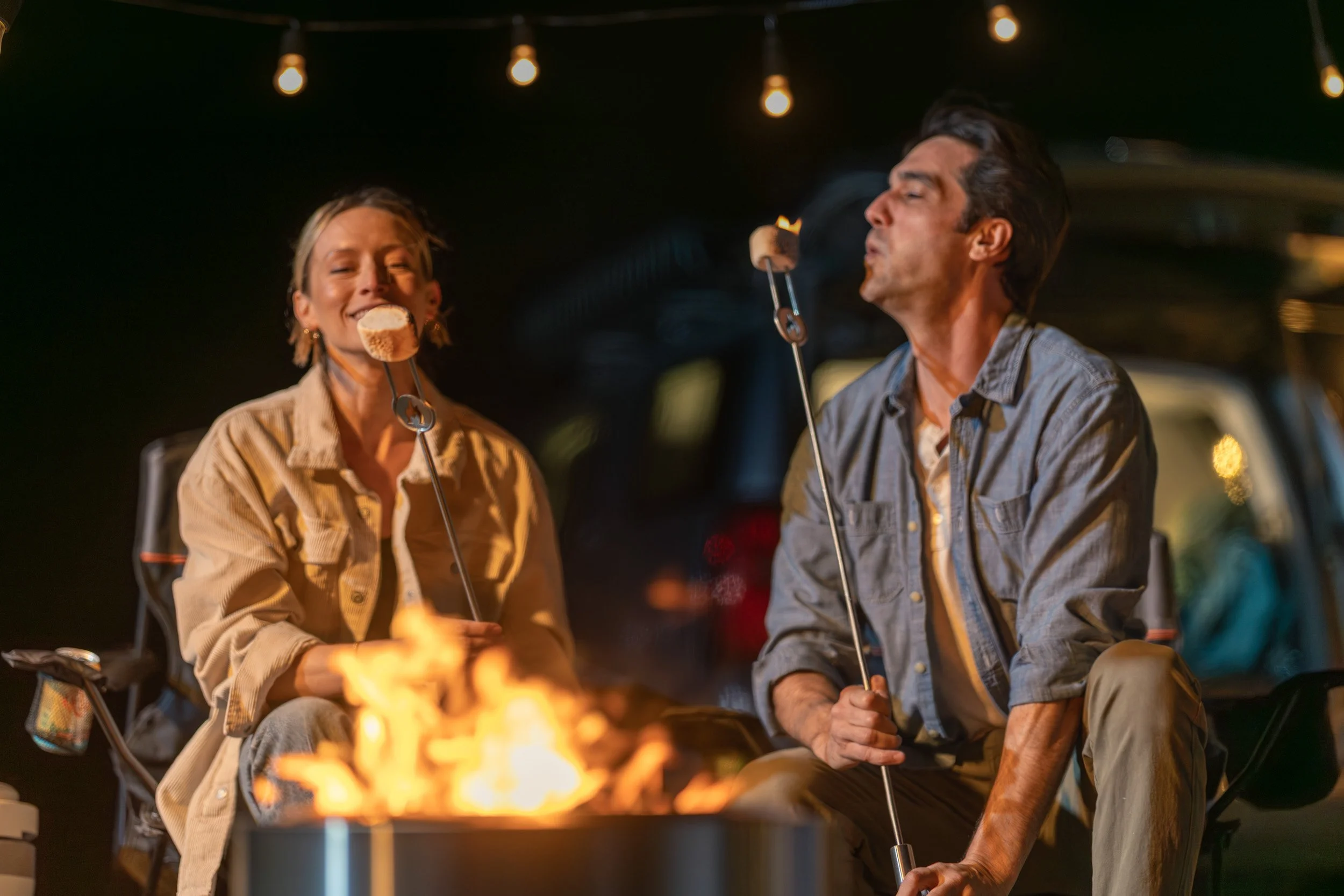 A man and woman enjoying marshmallows roasted over a campfire at night, with string lights above them.