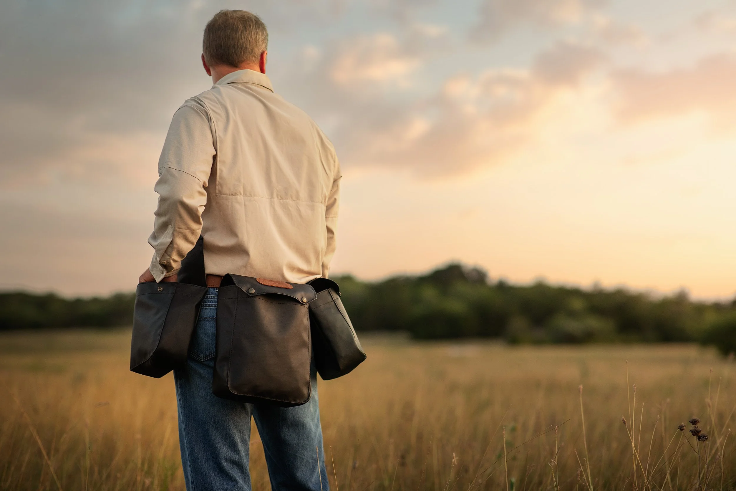 A man standing in a grassy field at sunset, viewed from behind, holding a black bag, wearing a beige jacket and blue jeans.