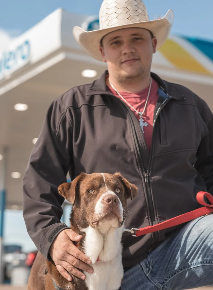 A man wearing a cowboy hat and a black jacket poses outdoors with a brown and white dog on a red leash, near a gas station.