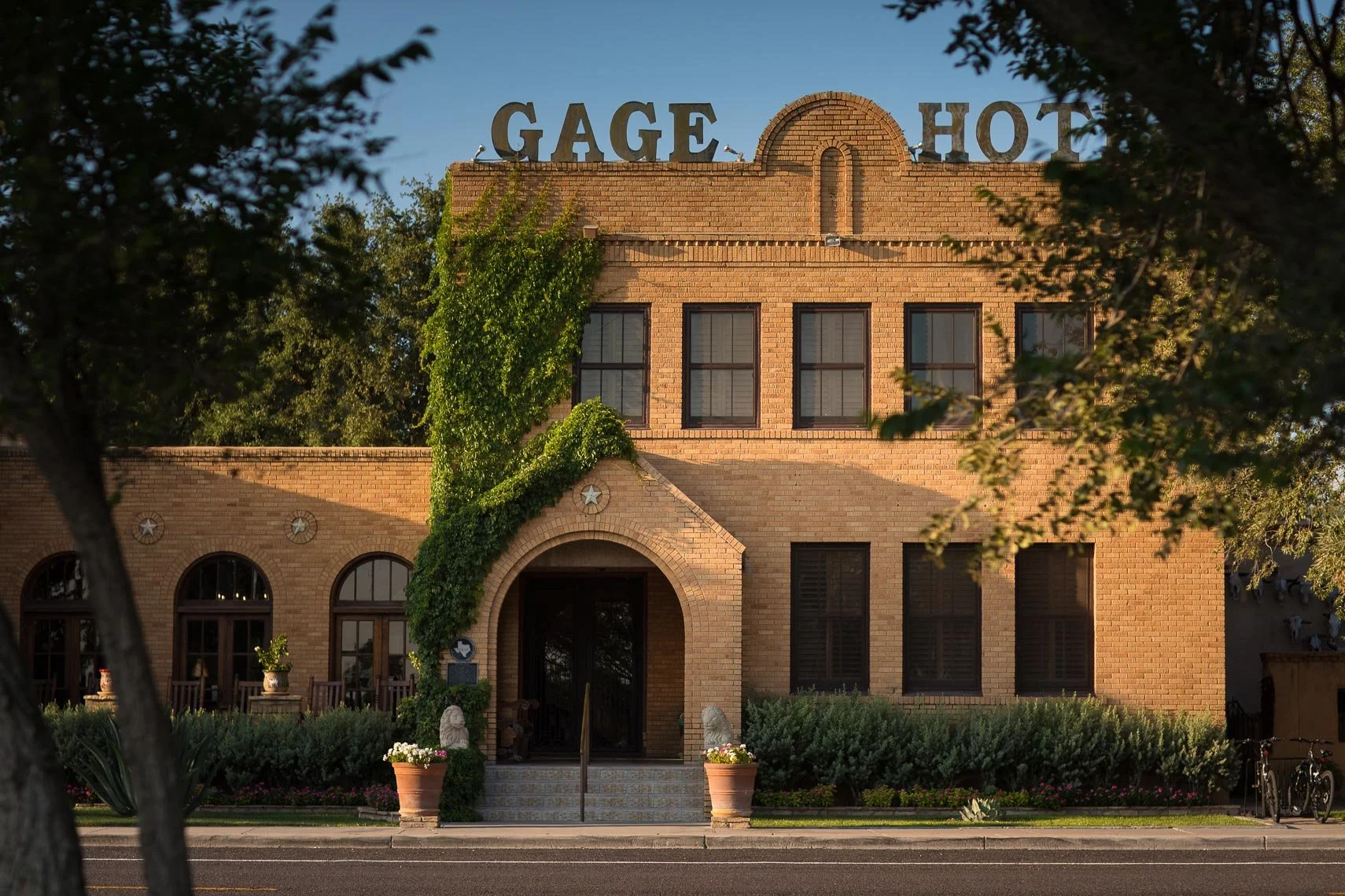 A brick building with the sign "GAGE HOTEL" on top, partially obscured by trees, with a central arched entrance and three windows above the doorway.