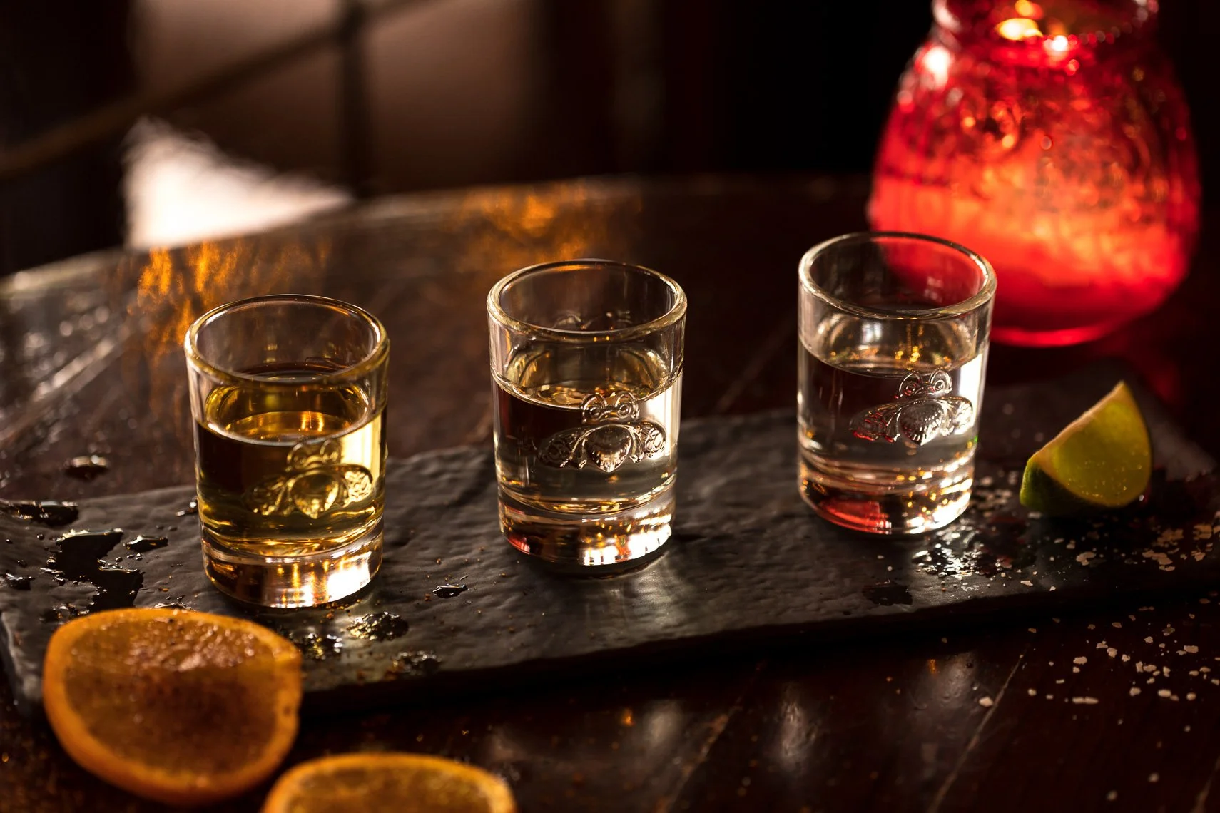 Three shot glasses filled with clear drinks with embedded teddy bear designs, placed on a dark stone serving board with orange slices and a lime wedge, in a dimly lit environment with a decorative red object in the background.