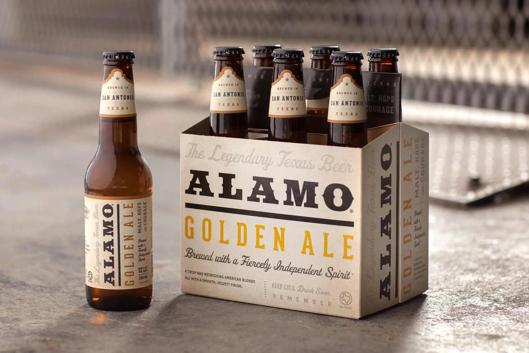 A six-pack of Alamo Golden Ale beer bottles with one bottle placed outside the pack on a surface, and a cardboard carrier holding remaining bottles, with background of a metal surface and blurred objects.