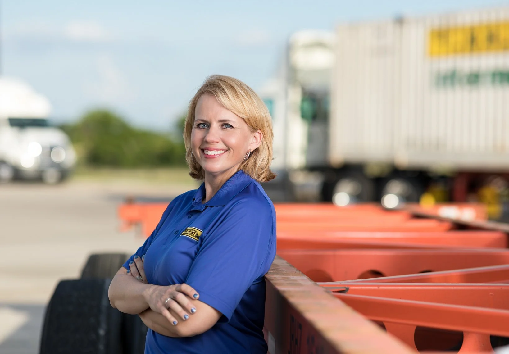 A smiling woman in a blue work shirt with a yellow logo, standing outdoors with her arms crossed, next to orange safety barriers, with trucks and a clear sky in the background.
