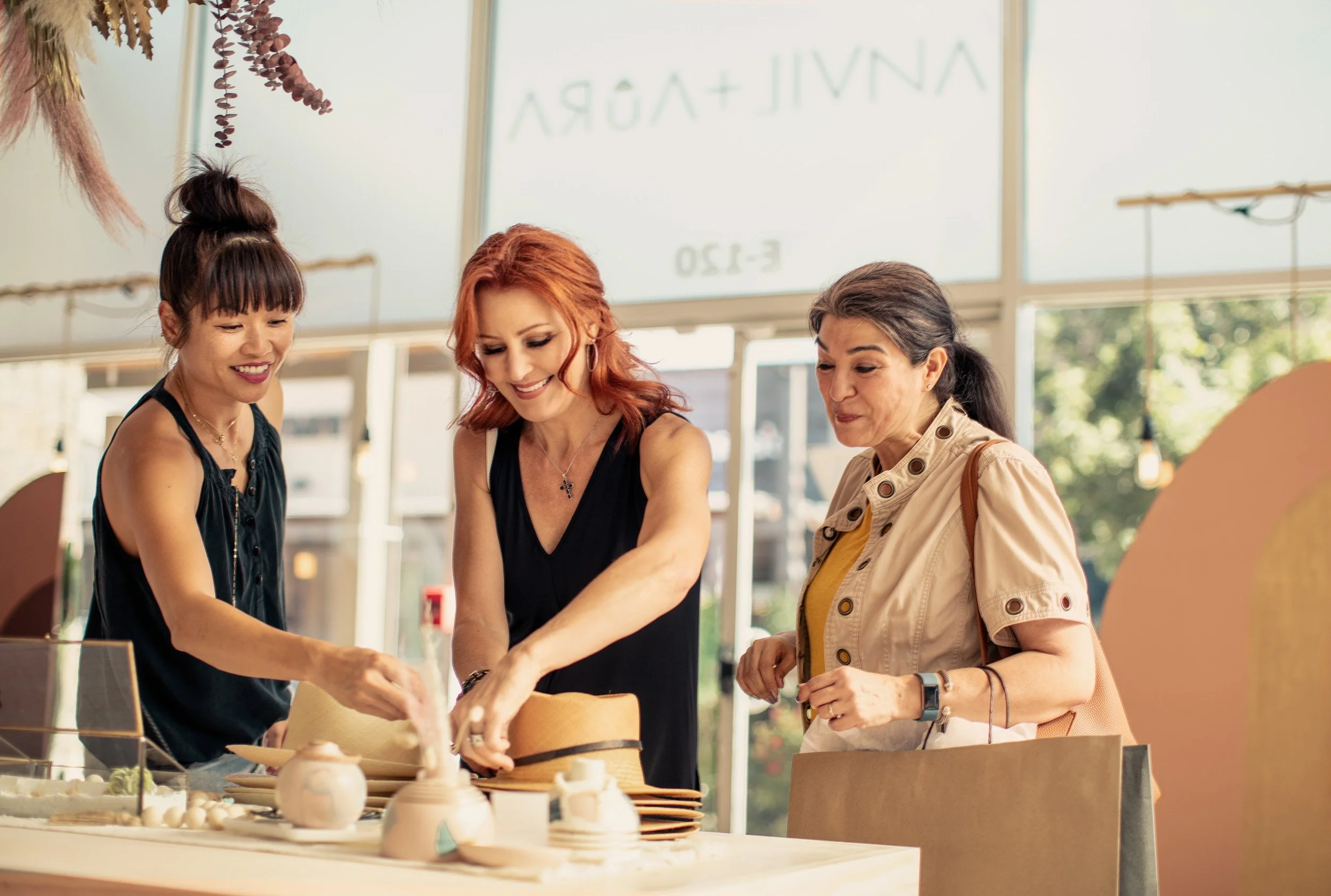 Three women looking at pottery on display in a bright, modern shop with a large window and greenery outside.