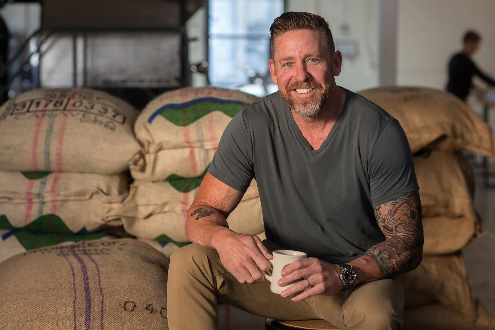 A man with a beard and tattoos, wearing a gray t-shirt, sitting and smiling while holding a white mug, with large sacks of coffee beans in the background.