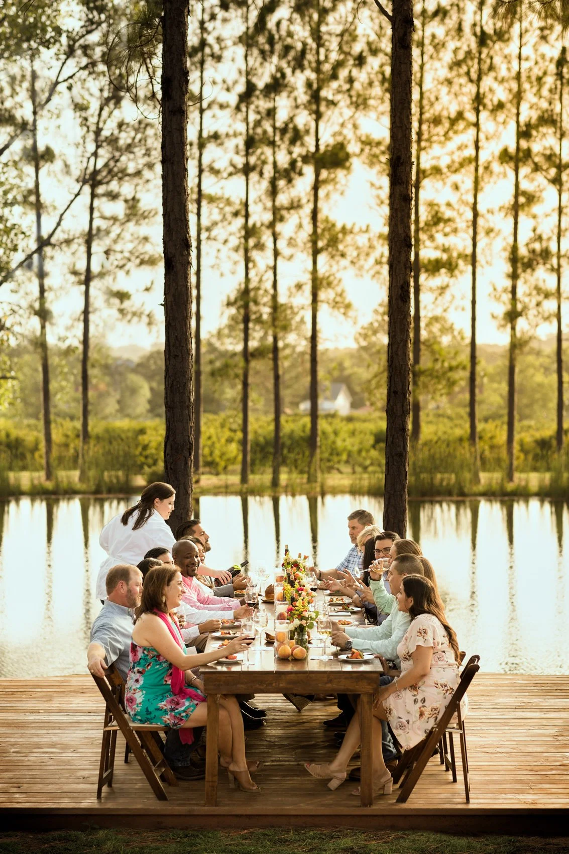 Group of people having dinner outdoors on a wooden deck by a lake in a forest at sunset, with a diverse mix of men and women seated at a long table decorated with flowers and fruits.