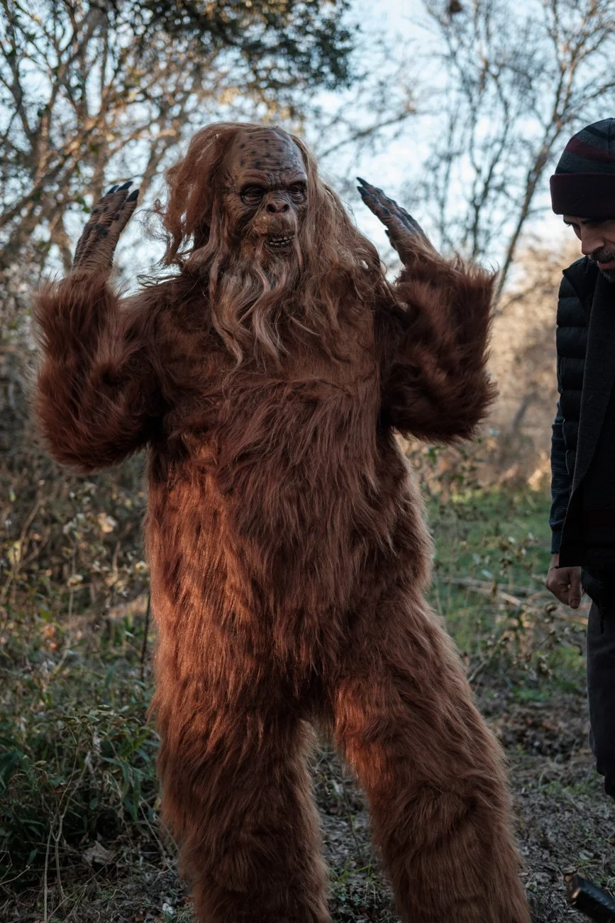 Person dressed in a Bigfoot costume with long hair, standing outdoors in a wooded area, with another person nearby.