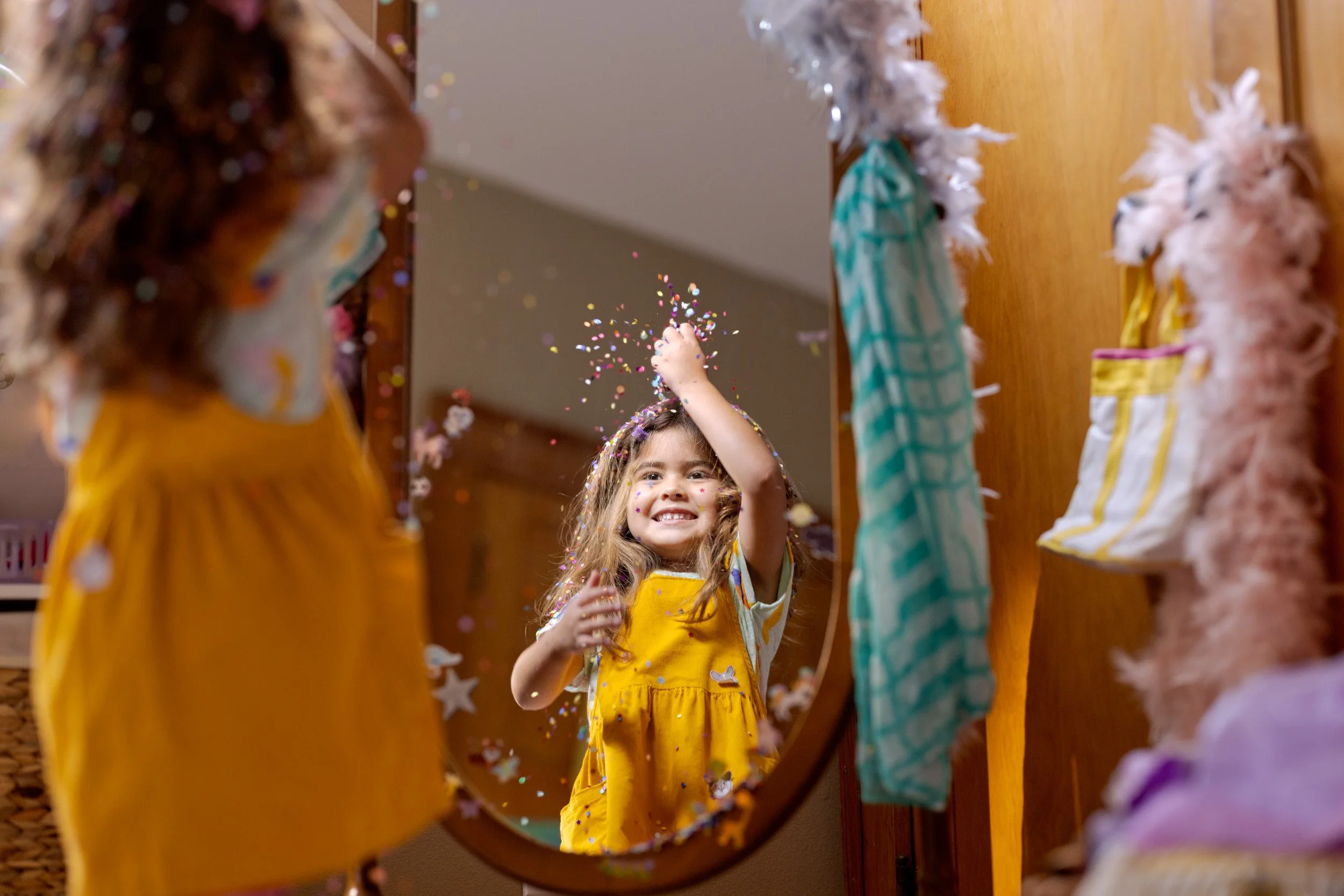 A young girl in a yellow dress with star patterns smiling and throwing colorful confetti while looking at her reflection in a mirror. The scene appears at a festive or party setting, with a wooden floor and colorful decorations around.