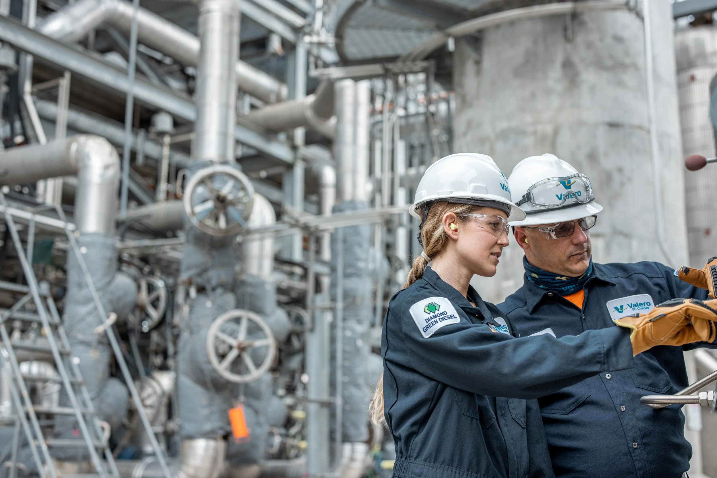 Two workers wearing hard hats, safety goggles, and navy uniforms inspecting equipment in an industrial plant with metal pipes and machinery.