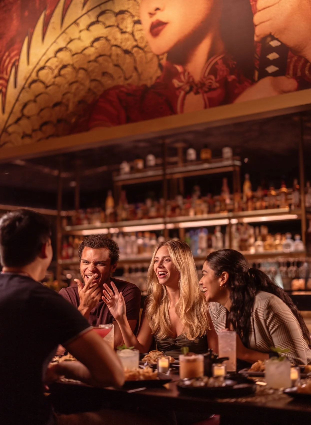 Group of four young adults, three women and one man, laughing and talking at a bar or restaurant, with various drinks and food on the table, in a warmly lit, lively atmosphere with a bar in the background and a large vintage portrait on the wall abov