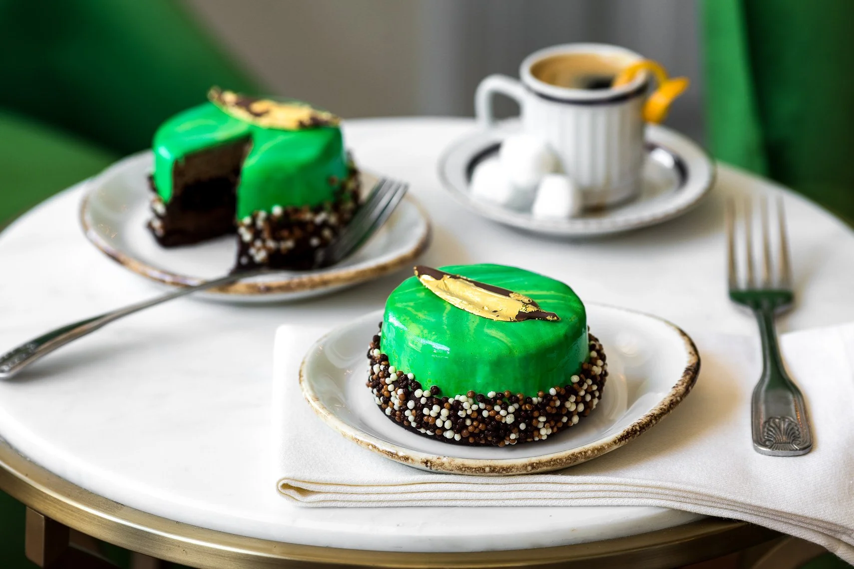 Close-up of a round, green glazed dessert topped with a gold leaf, surrounded by small white, brown, and black sprinkles, served on a white plate with a fork nearby. In the background, a slice of the same dessert and a cup of coffee with marshmallows