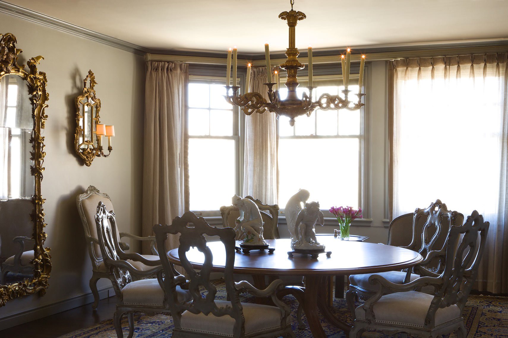A vintage dining room with a wooden table, ornate chairs, porcelain figurines, a vase of pink flowers, a large gilded mirror, and a chandelier hanging from the ceiling near large windows with curtains.
