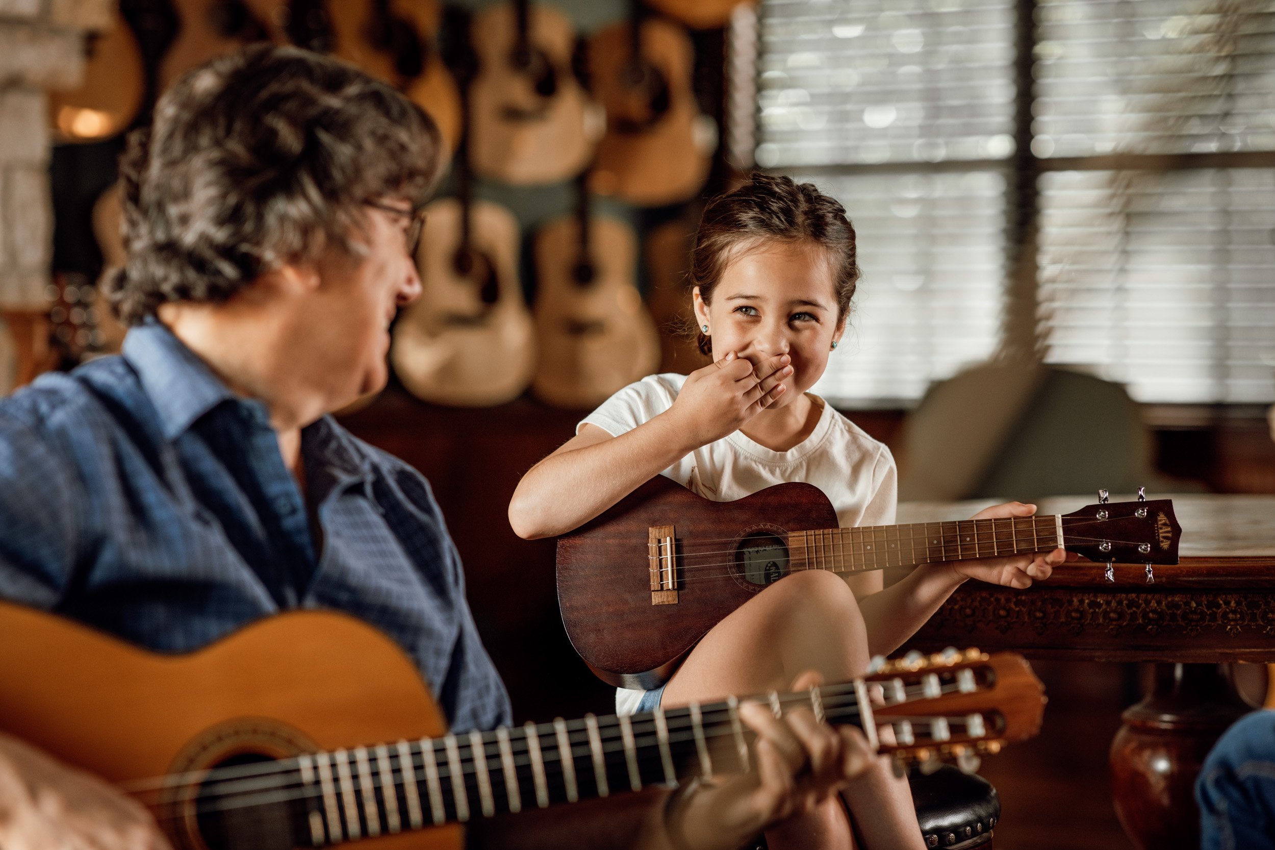 A young girl happily covering her mouth with her hand while holding a guitar, sitting next to an older woman playing a guitar, in a room with a wall of acoustic guitars in the background.