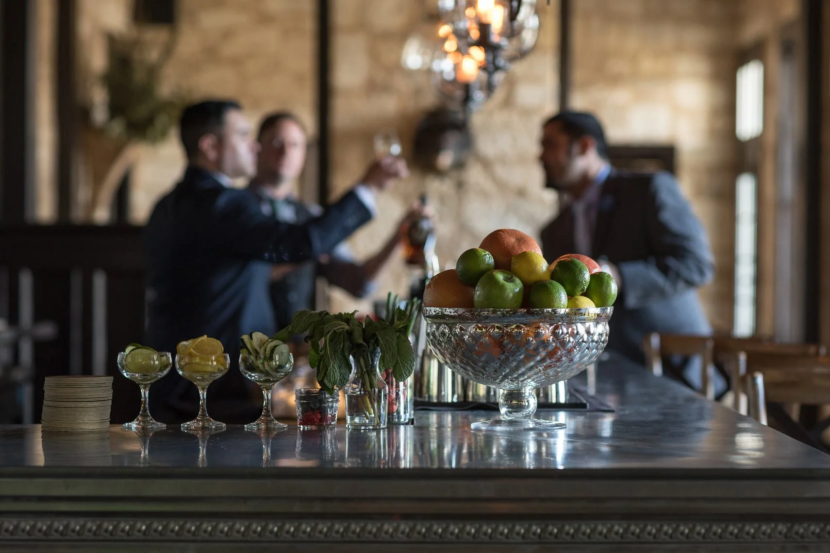 A bar or reception area with a large glass bowl filled with apples, limes, and peaches on a marble countertop, with small drinks, mint leaves, and utensils in the foreground, and three men in suits talking in the background inside a rustic brick-wall