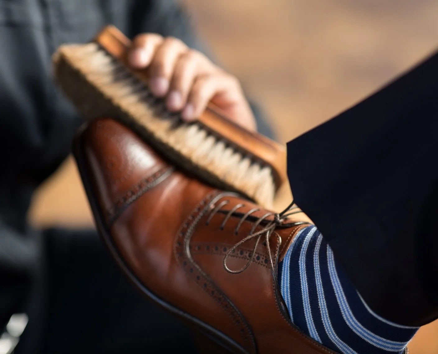 Close-up of a man wearing polished brown leather dress shoes with dark laces, striped dark blue and white socks, and black trousers, resting his foot on a wooden surface. In the background, a hand is holding a small wooden percussion instrument with 