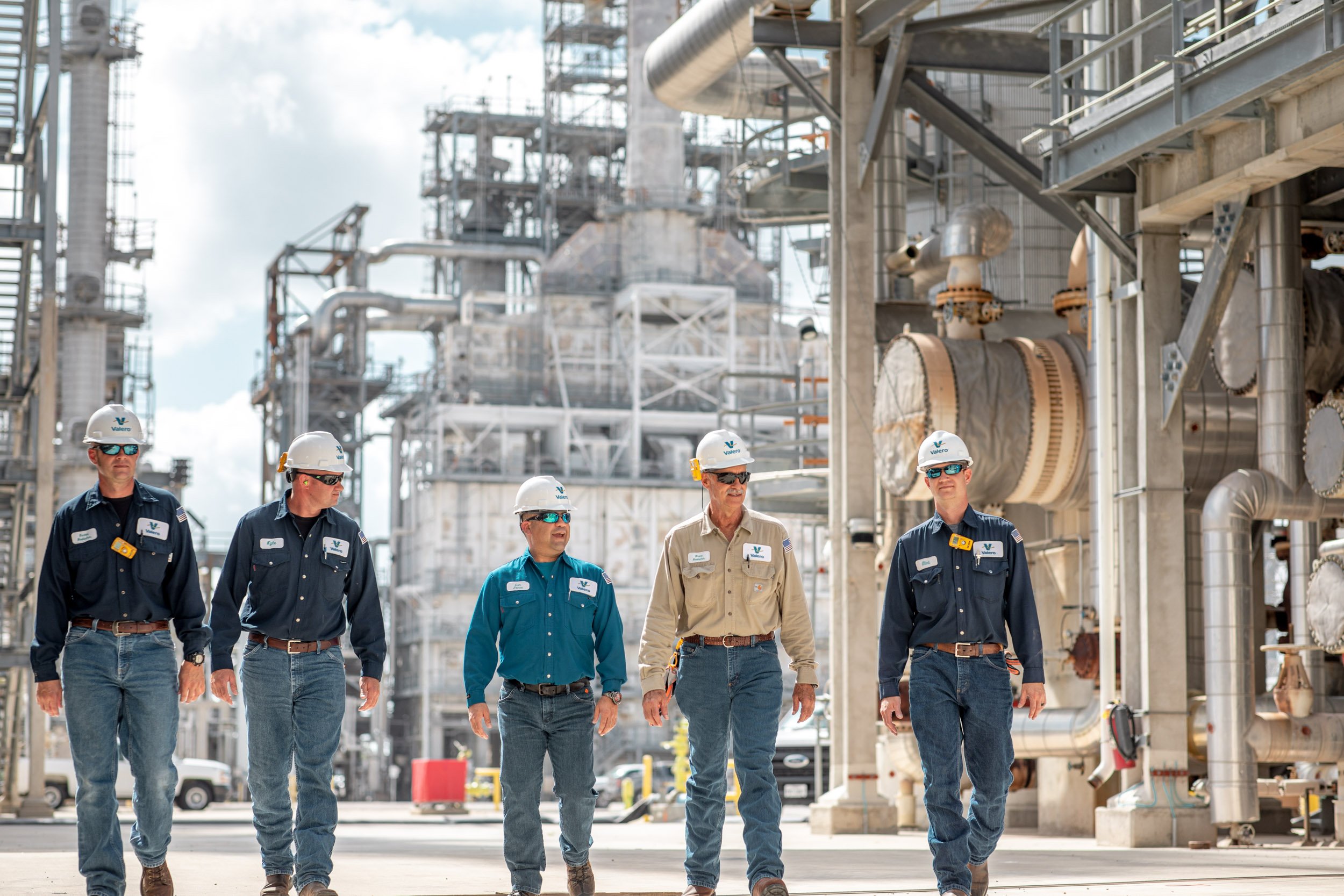 Group of five industrial workers walking through factory with machinery and pipes in background. All are wearing safety helmets, some have sunglasses, and their shirts have company logos.