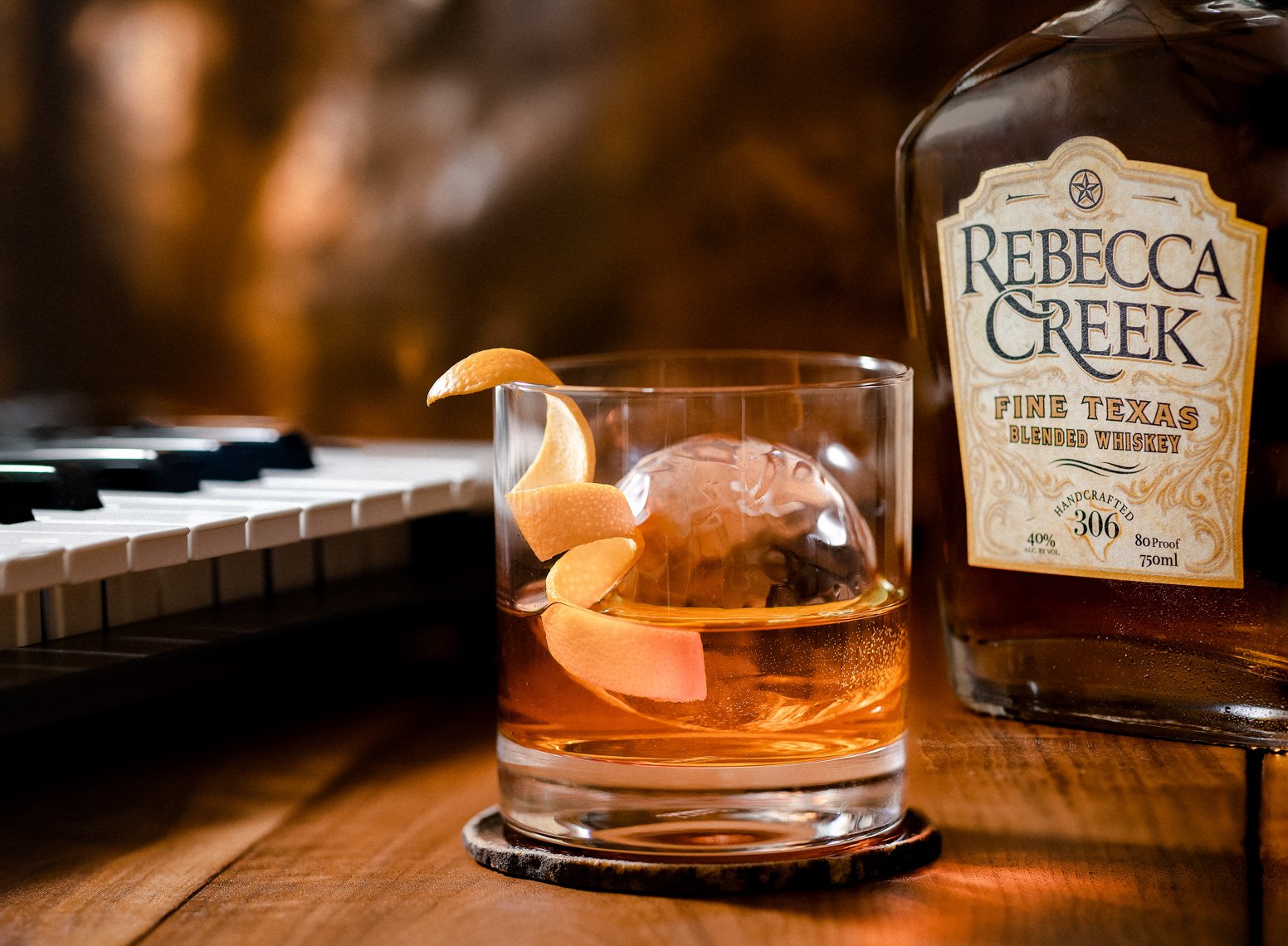 A glass of whiskey with lemon twist on a wooden table, a bottle of Rebecca Creek blended whiskey nearby, and a keyboard in the background.