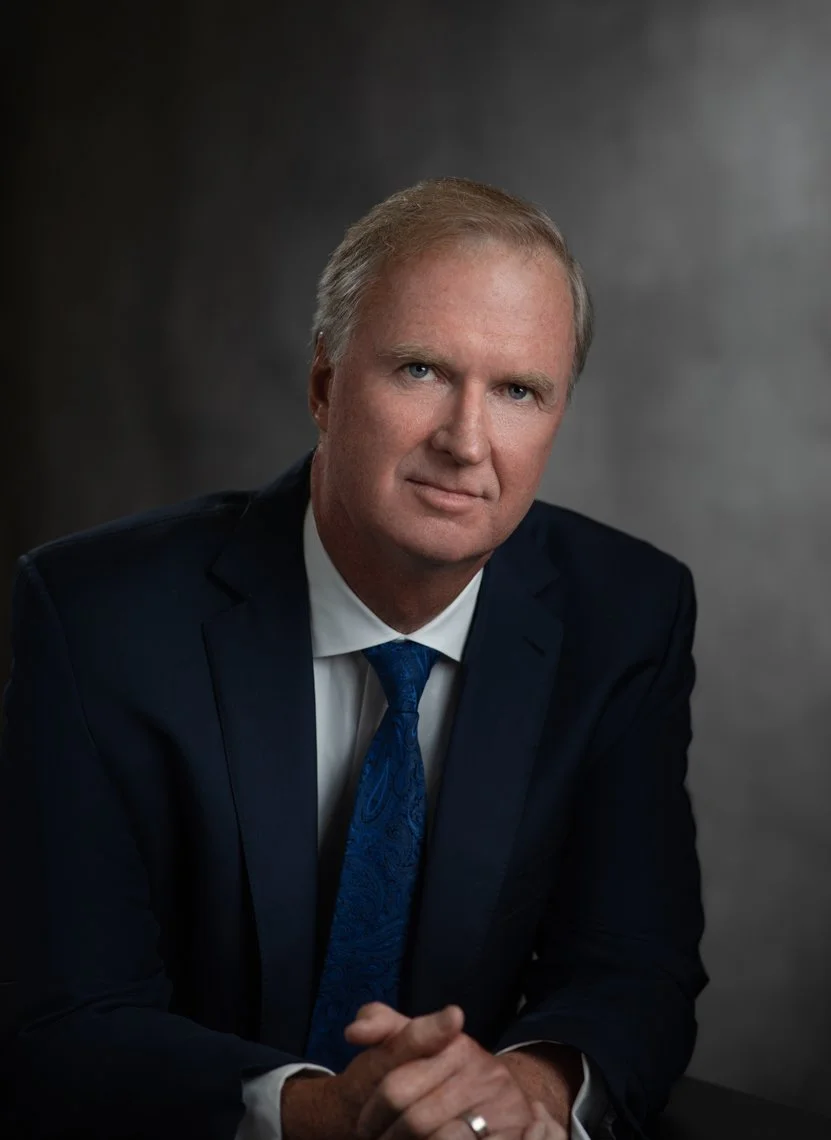 A middle-aged man with light skin, blonde hair, wearing a dark suit, white shirt, and blue tie, sitting with hands clasped in front of a dark gray background.