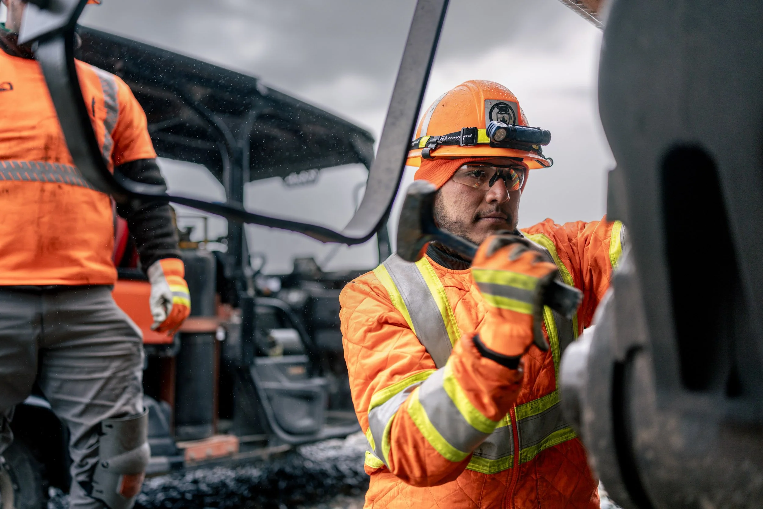 A worker in a safety orange jacket and hard hat adjusting equipment near a construction vehicle on a cloudy day.