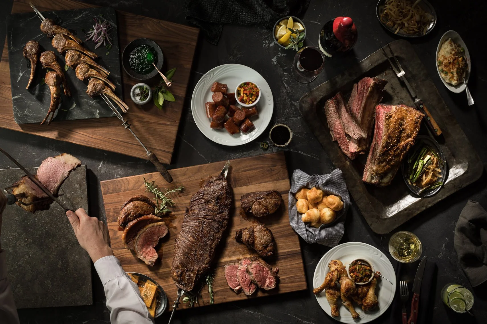 A table with various cuts of cooked and grilled meats, including beef steaks, ribs, and lamb chops, along with side dishes and sauces, set on a dark surface.