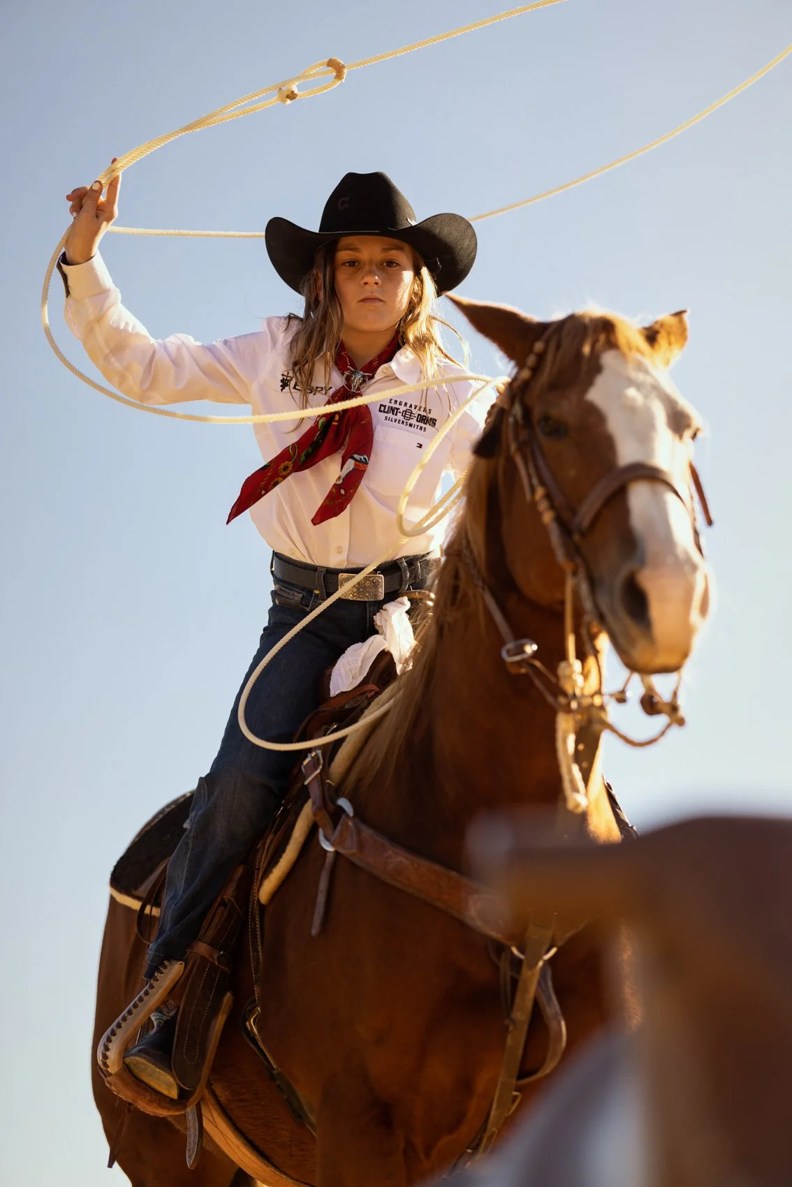 A young girl dressed as a cowboy riding a brown horse, holding a lasso, wearing a black cowboy hat, white shirt, red bandana, and jeans, against a clear blue sky.