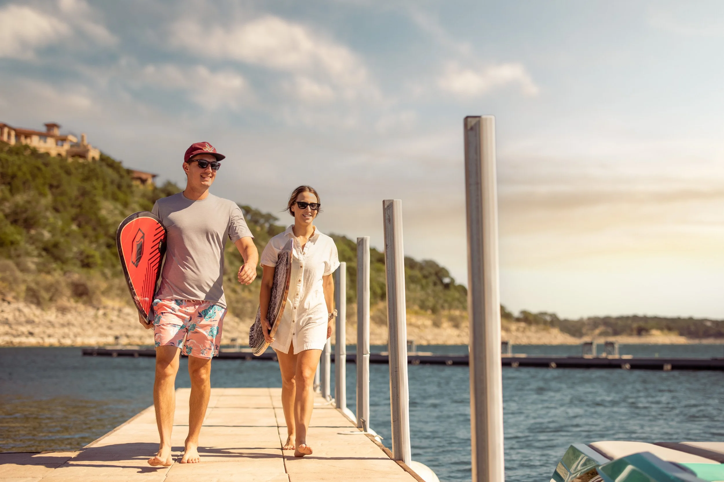 A man and woman walking barefoot on a dock by the water, carrying surfboards, smiling, with a house on a hill and a cloudy sky in the background.