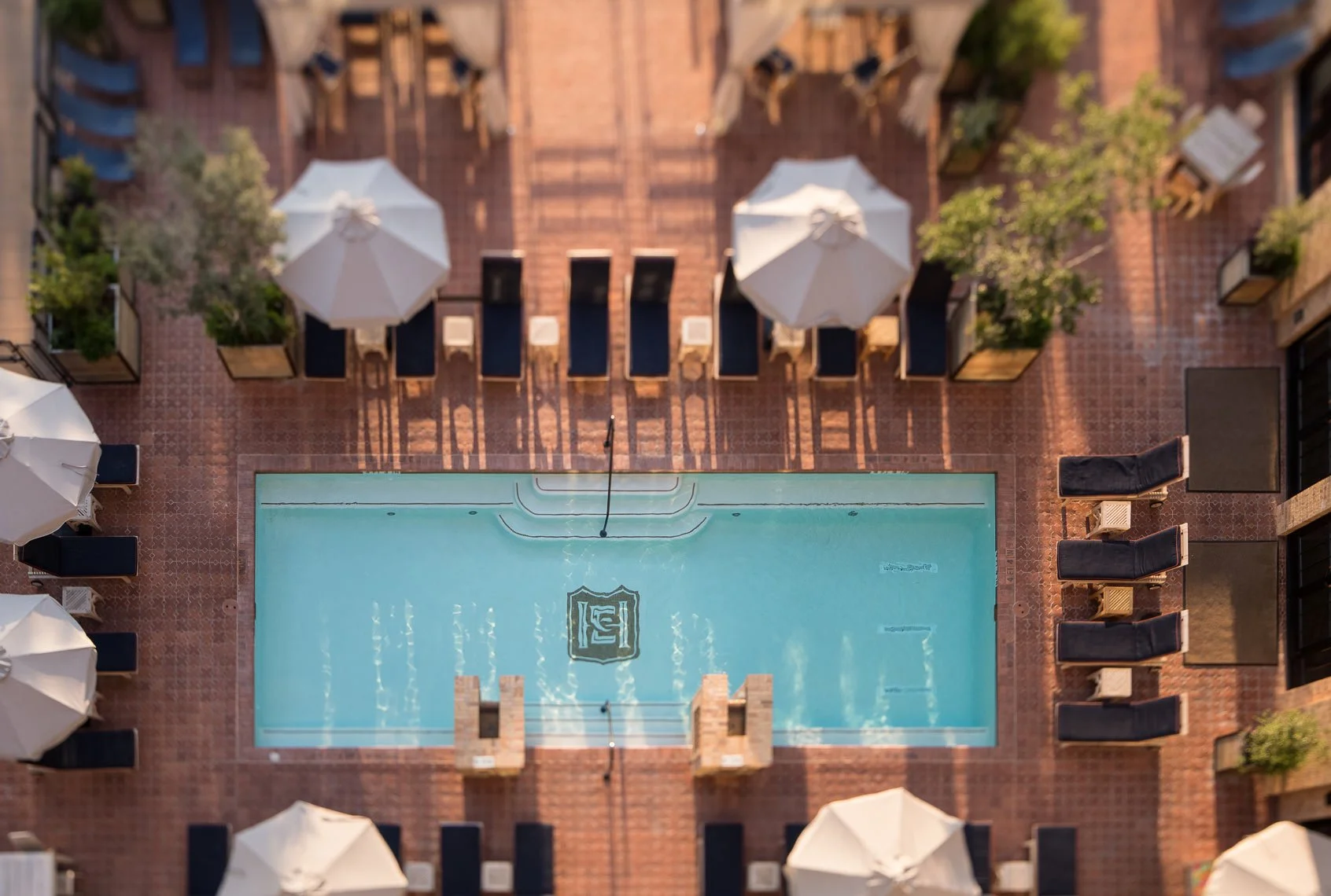 A rooftop swimming pool area with lounge chairs, umbrellas, greenery, and an American flag painted on the pool bottom, viewed from above.