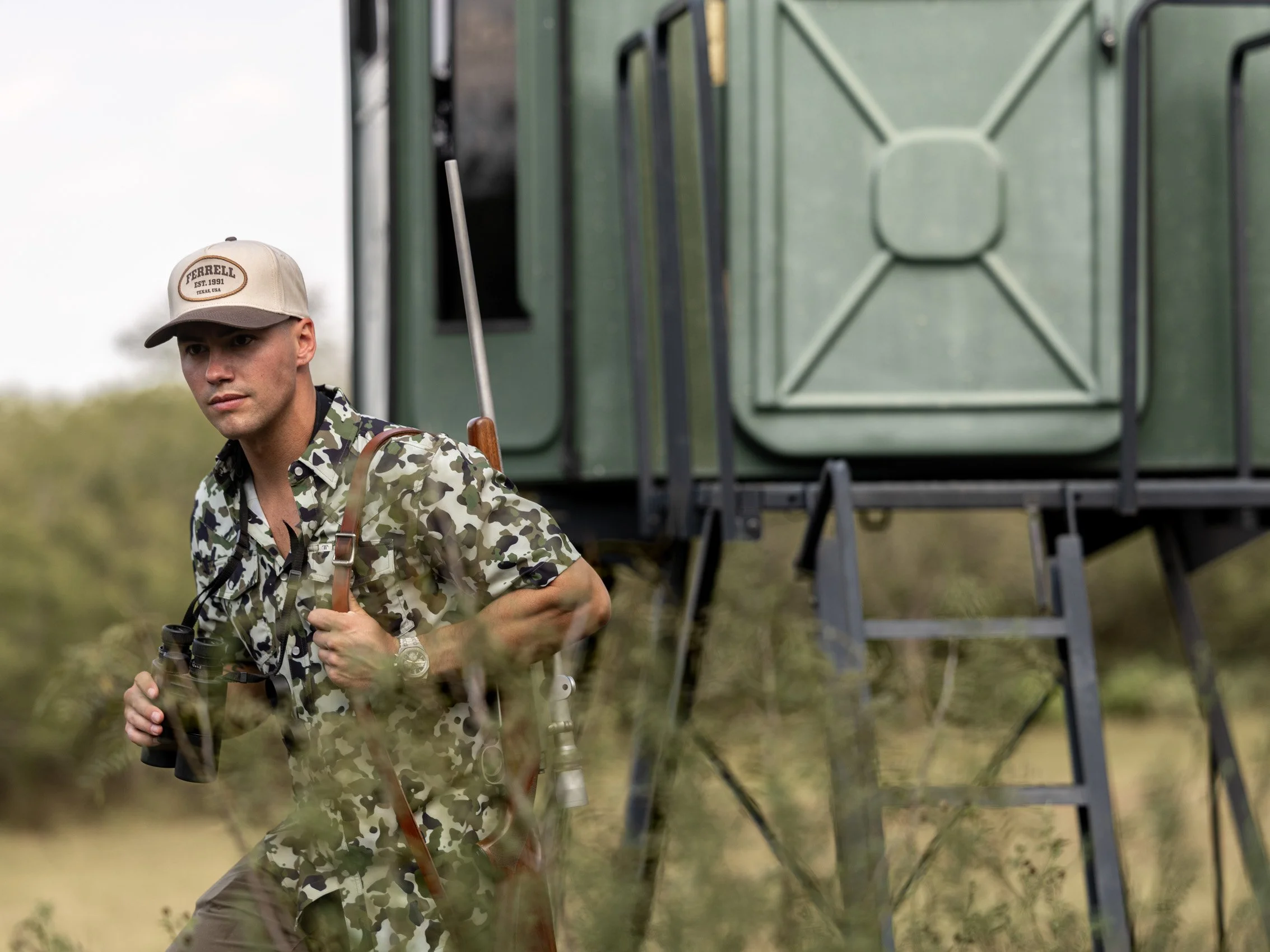 A man in camouflage clothing and a beige cap outdoors, holding binoculars with a green blind in the background.