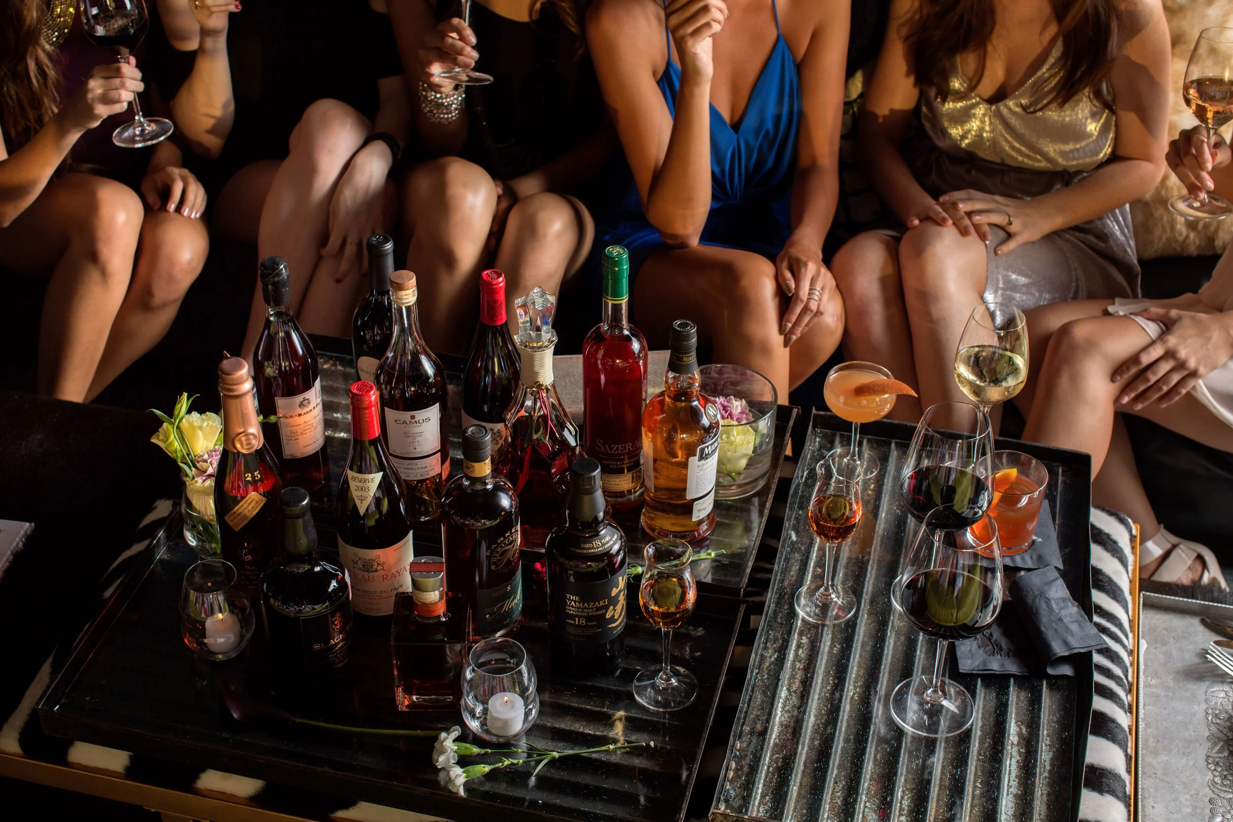 Group of women dressed in party attire sitting around a table with wine bottles, glasses, and cocktails at a celebration or party.