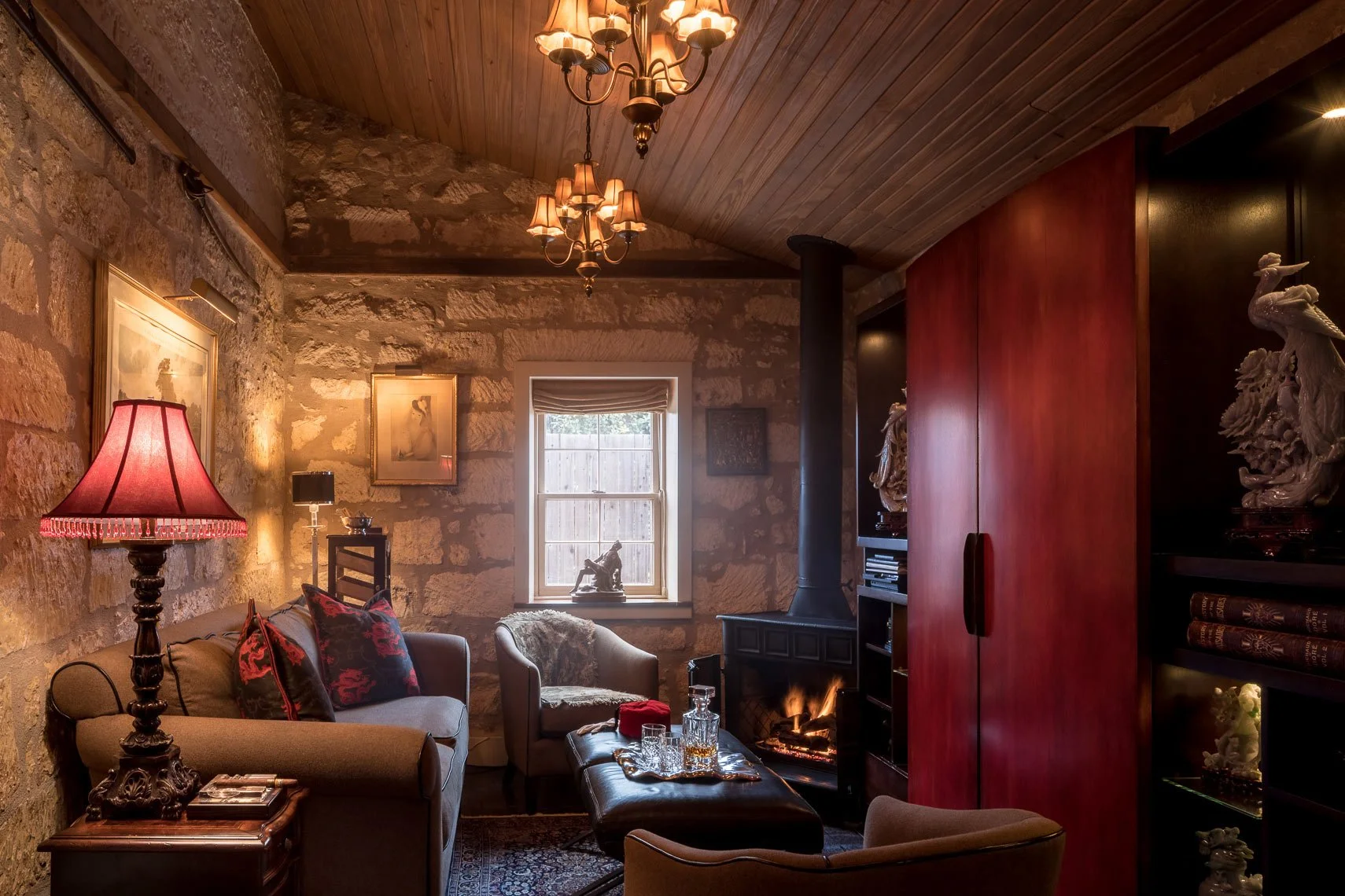 Cozy living room with stone walls, a wood-paneled ceiling, and a fireplace. The room features a red' cabinet, various chairs, a table with glassware, a red lampshade, and decorative items.
