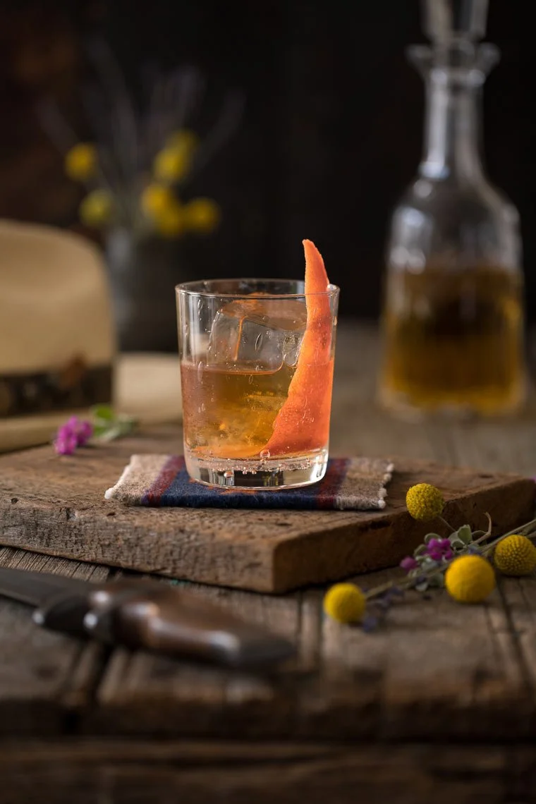 A glass of whiskey with ice and a twist of orange peel garnishing it, placed on a rustic wooden table with a small cloth underneath, surrounded by yellow and purple flowers, with a bottle of whiskey and other items blurred in the background.