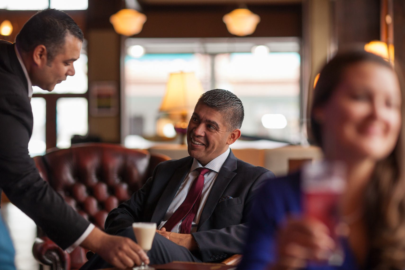 Two men in suits shaking hands in a restaurant or cafe, with a woman in the foreground smiling and holding a drink. The background shows warm lighting, lamps, and windows.