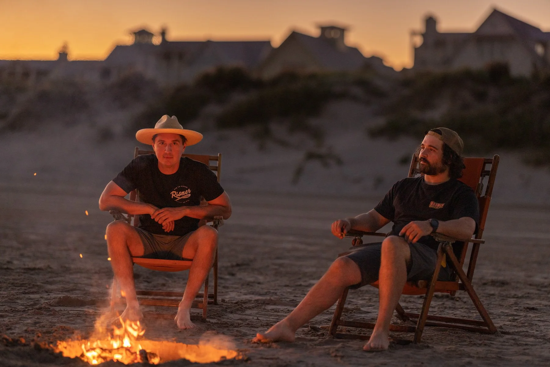 Two men sitting in chairs around a campfire on a beach at dusk, with houses in the background.