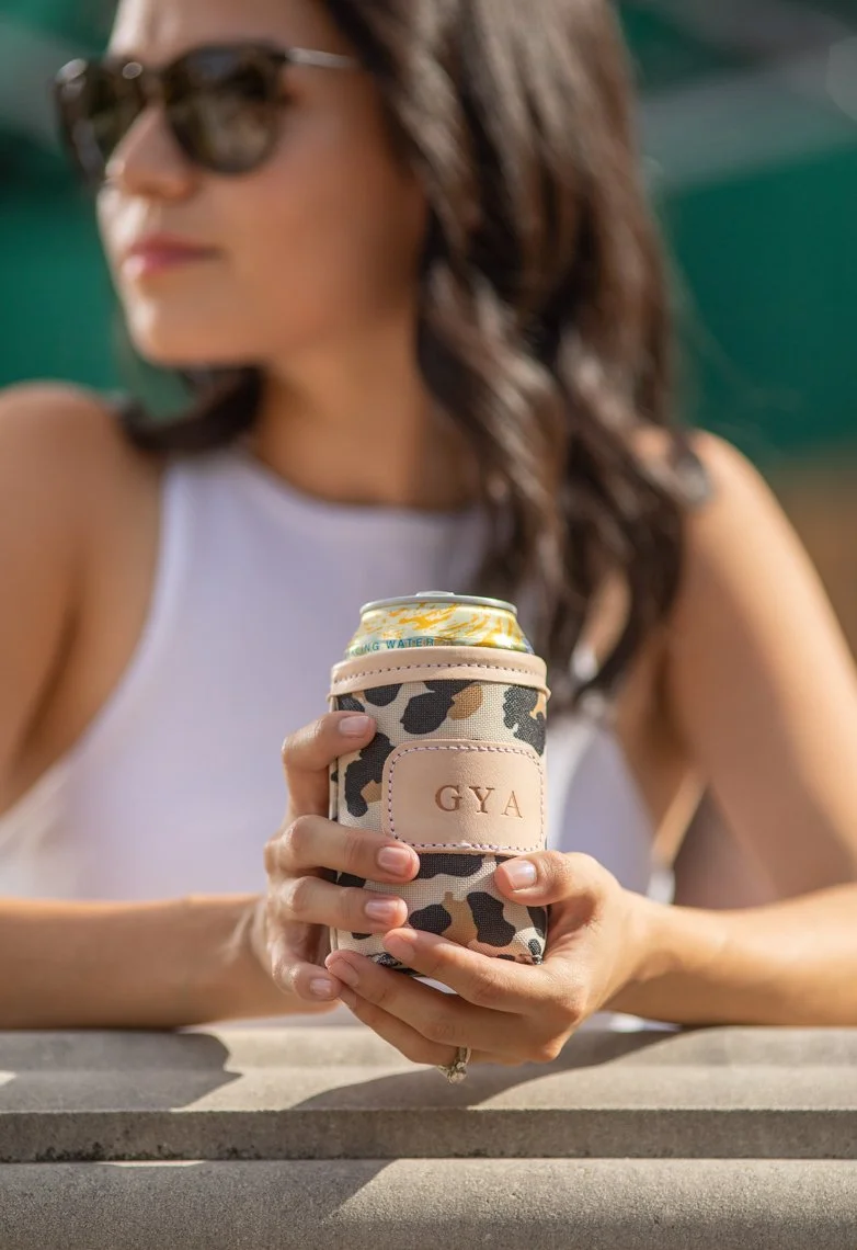 A woman with long brown hair, wearing sunglasses and a white sleeveless top, holding a can in a leopard-print can holder with the initials 'GYA' embossed on it. She is sitting outdoors during the daytime.