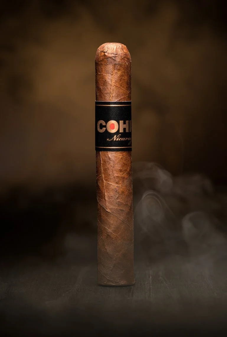A cigar standing upright on a dark surface with a blurred dark background, emitting a small amount of smoke.