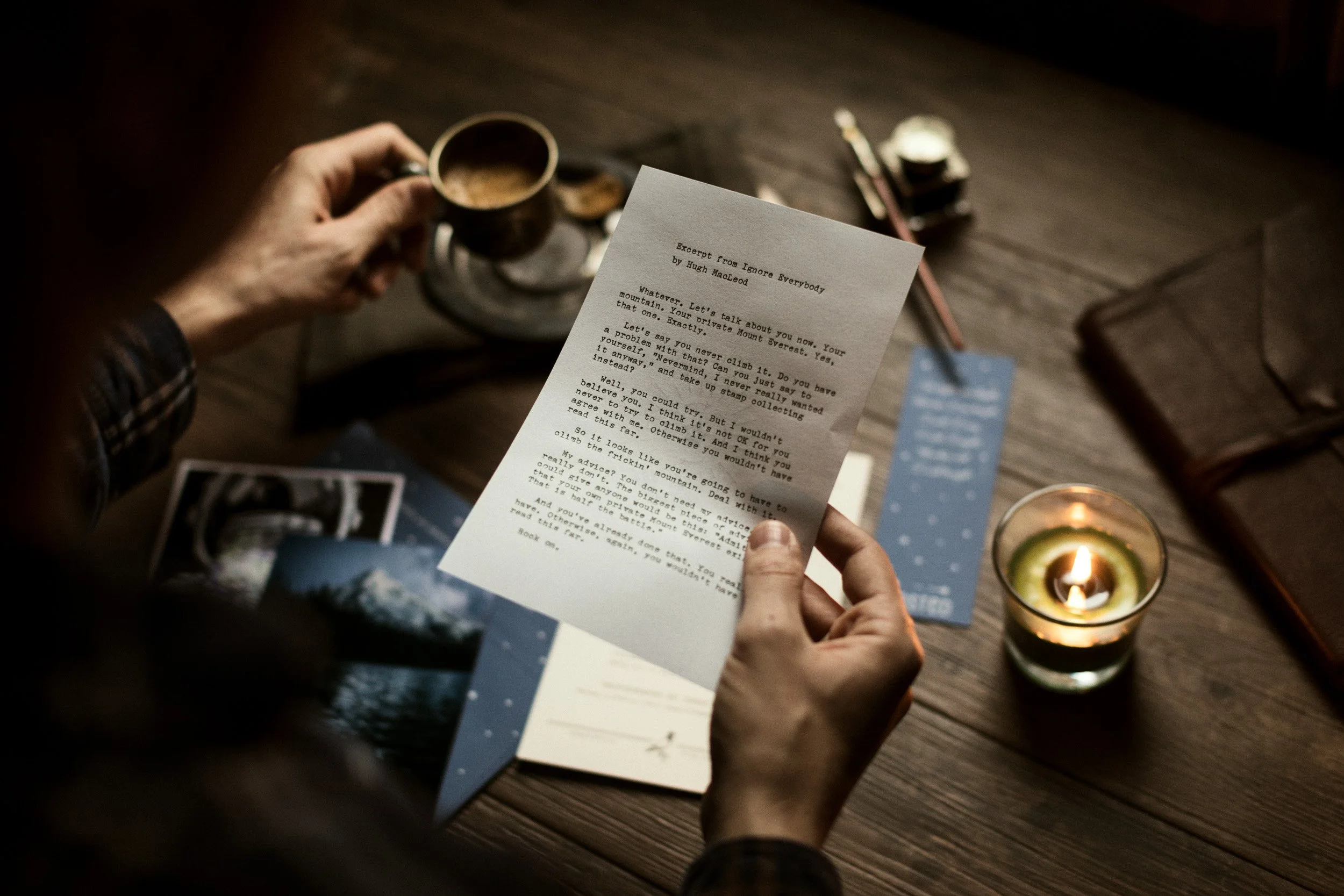 Person holding a printed excerpt with a coffee mug and lit candle on a wooden table.