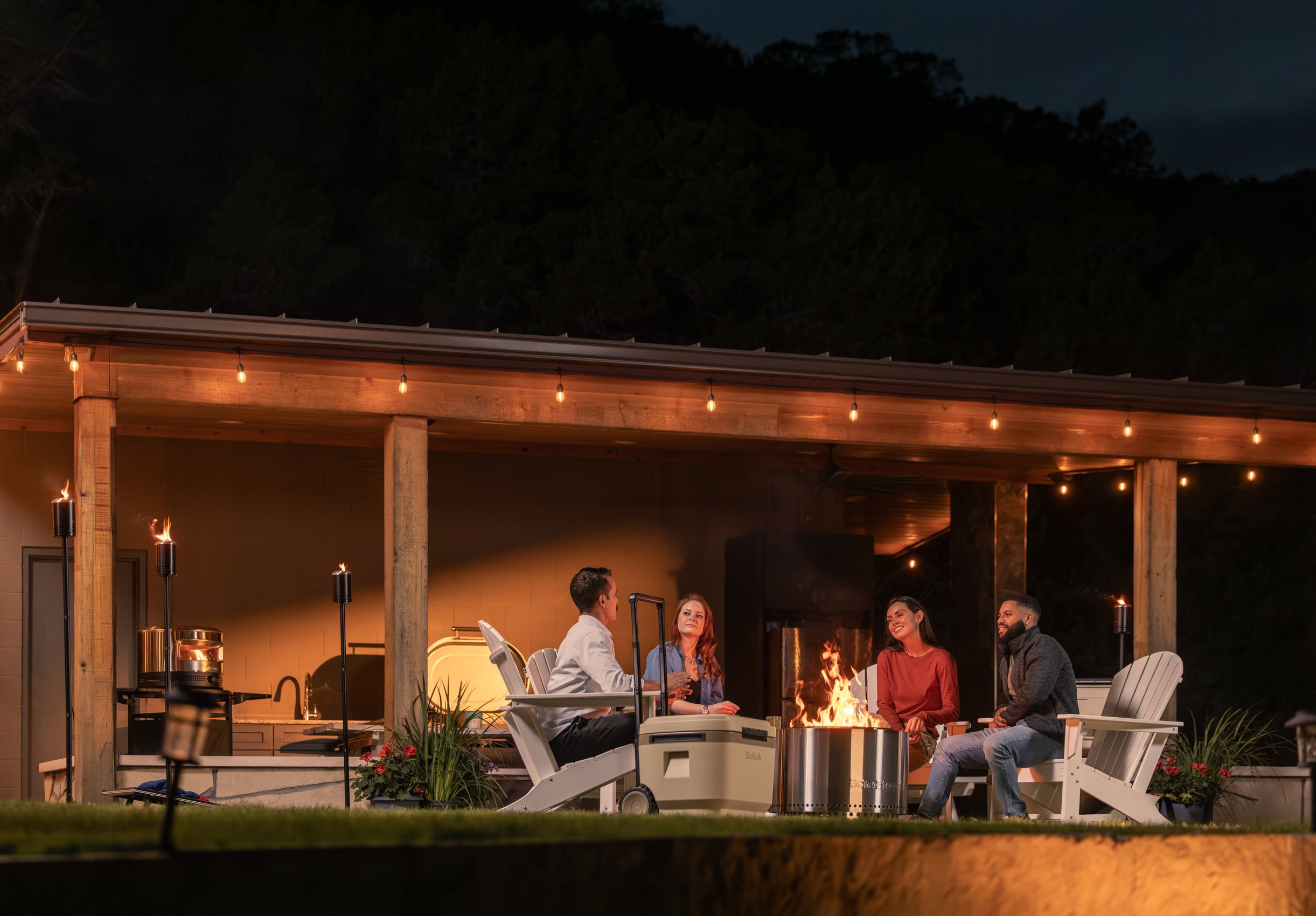 A group of people sitting around a fire pit on a covered patio at night, engaging in conversation. String lights are hanging above, and the patio has a warm, inviting atmosphere with trees and mountains visible in the background.