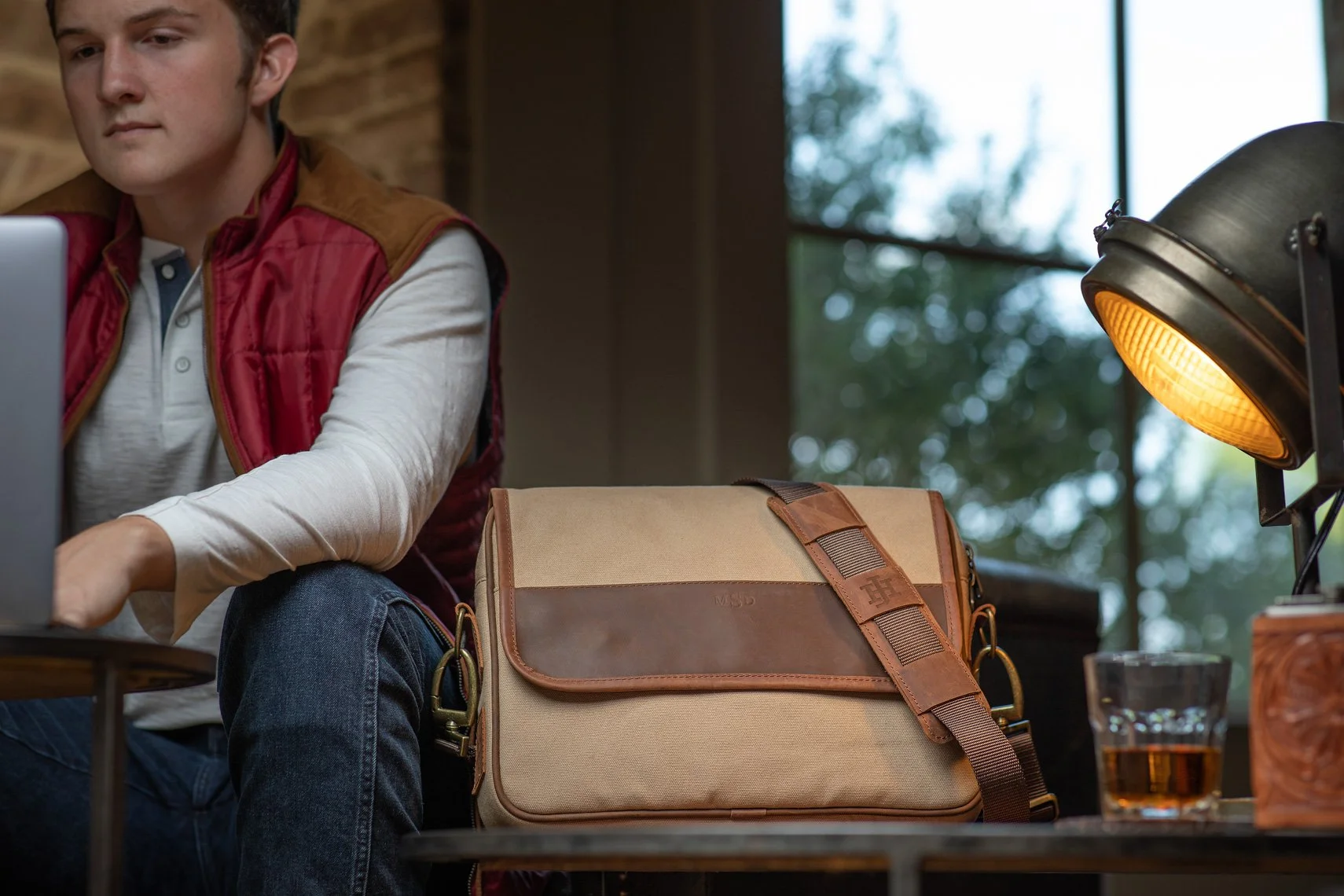 A man wearing a red vest and white shirt working on a laptop at a table. A beige and brown messenger bag is on the table near a desk lamp with warm lighting, and a glass of water is also visible. Large windows with blurred outdoor scenery are in the 