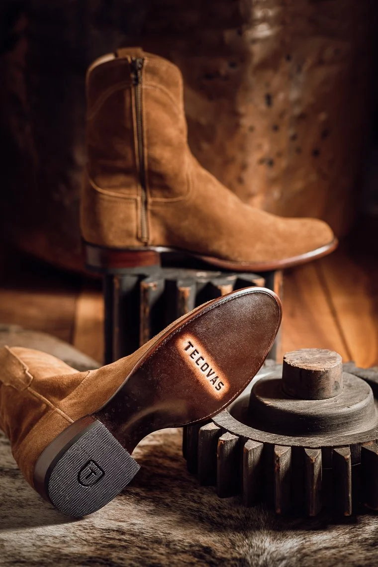 A tan suede ankle boot with a zipper, positioned on a wooden gear-shaped object, with a blurry background of rustic wood and leather textures.