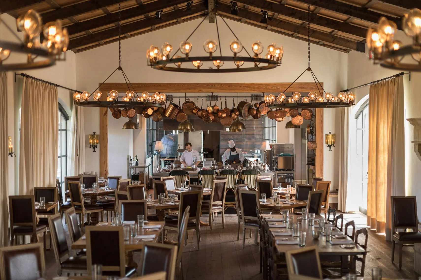 Elegant restaurant interior with wooden tables and chairs, chandeliers and copper pots hanging overhead, large windows with curtains, and chefs preparing food in the open kitchen area.