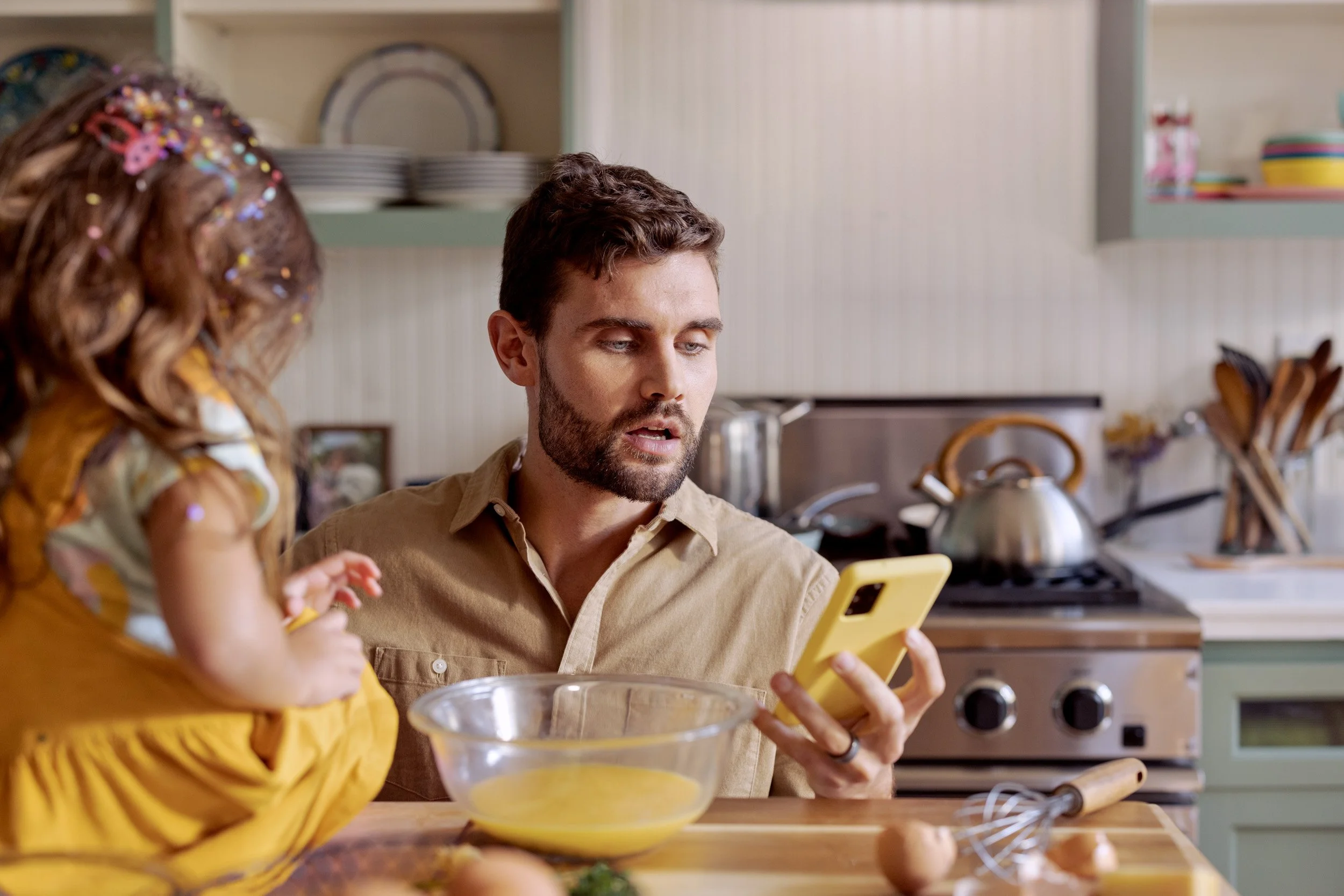 A man and a young girl in a kitchen looking at a smartphone while preparing food. The girl is wearing a yellow dress with colorful sprinkles in her hair, and the man is wearing a tan button-up shirt.